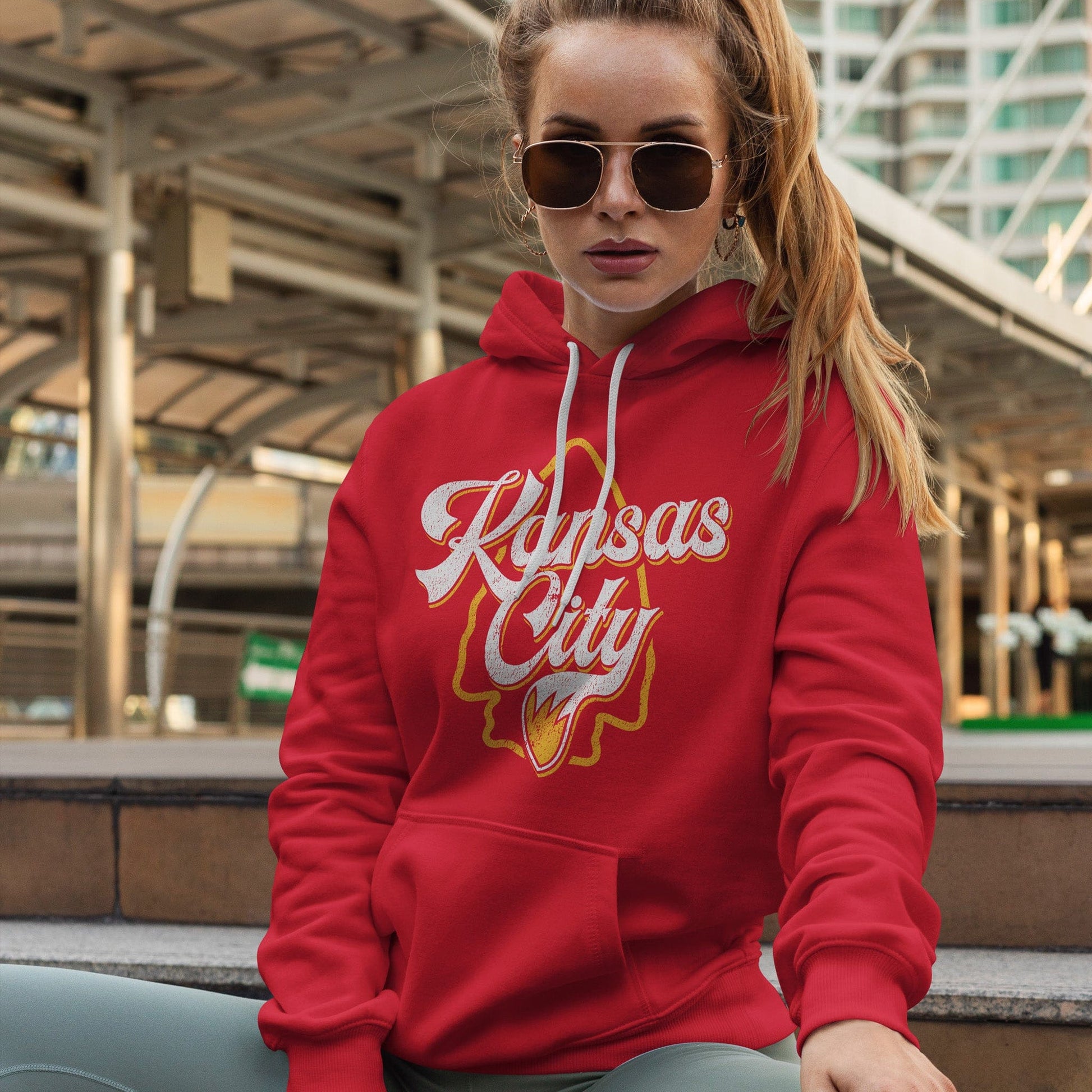 KC Swag | Kansas City Chiefs white/gold KC WOLFTAIL on red sponge-fleece pullover hoodie worn by female model sitting on steps in front of a downtown building