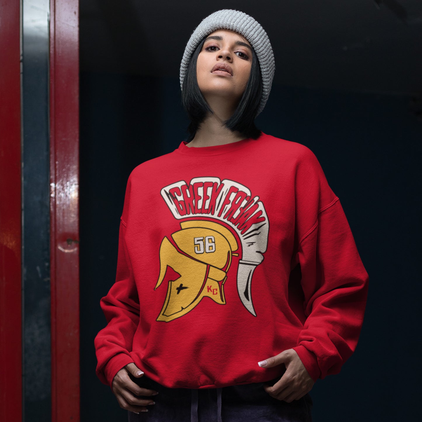 KC Swag Kansas City Chiefs White & Gold Greek Freak (with Trojan Helmet) on a Red Crewneck Sweatshirt worn by beaning wearing female model standing under a white light outside of a red door