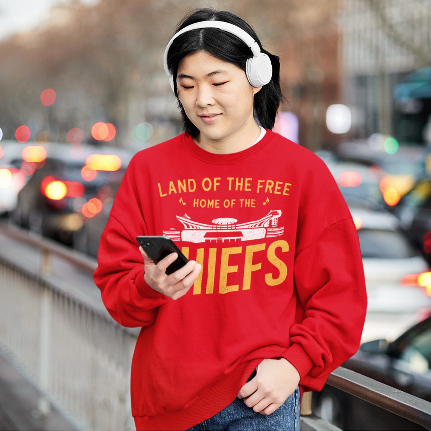 KC Swag Kansas City Chiefs Distressed White & Gold Home Of The Chiefs on a Red Crewneck Sweatshirt worn by a female model wwearing headphones on a busy downtown street
