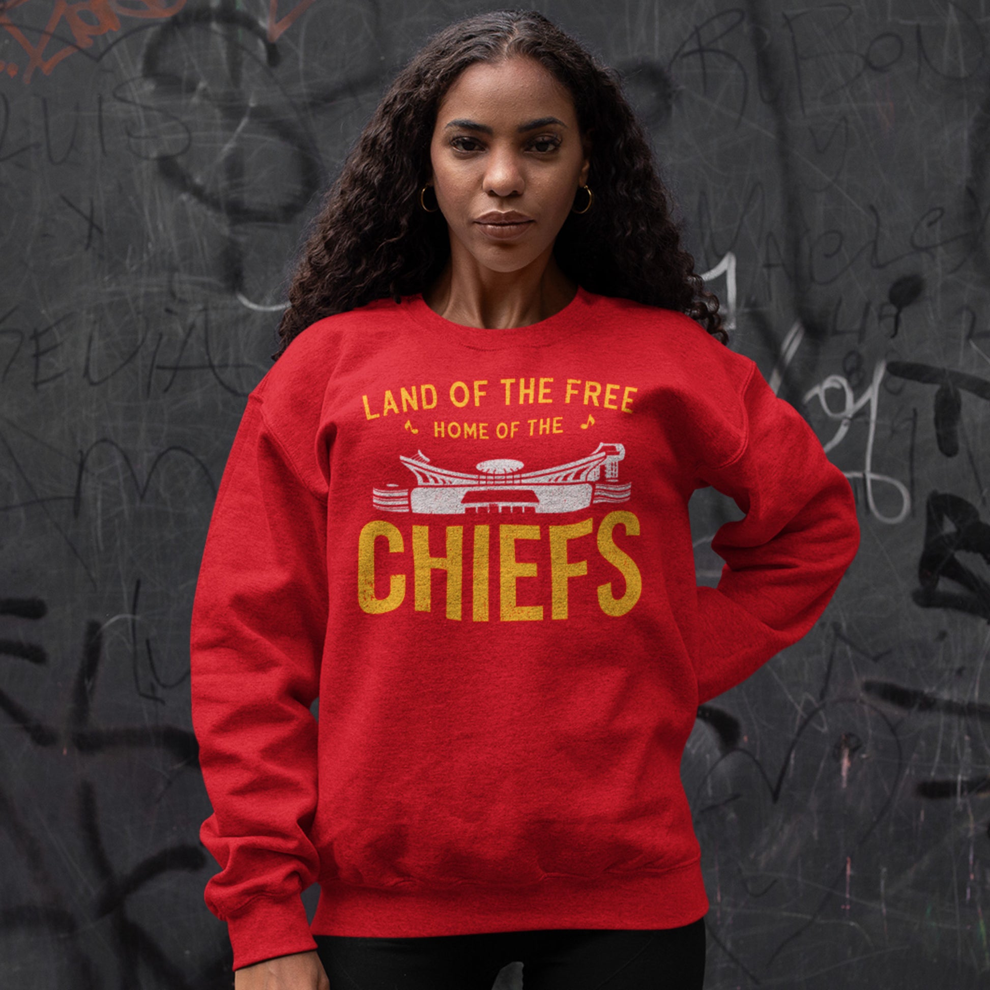 KC Swag Kansas City Chiefs Distressed White & Gold Home Of The Chiefs on a Red Crewneck Sweatshirt worn by female model standing in front of a dark gray graffitied wall