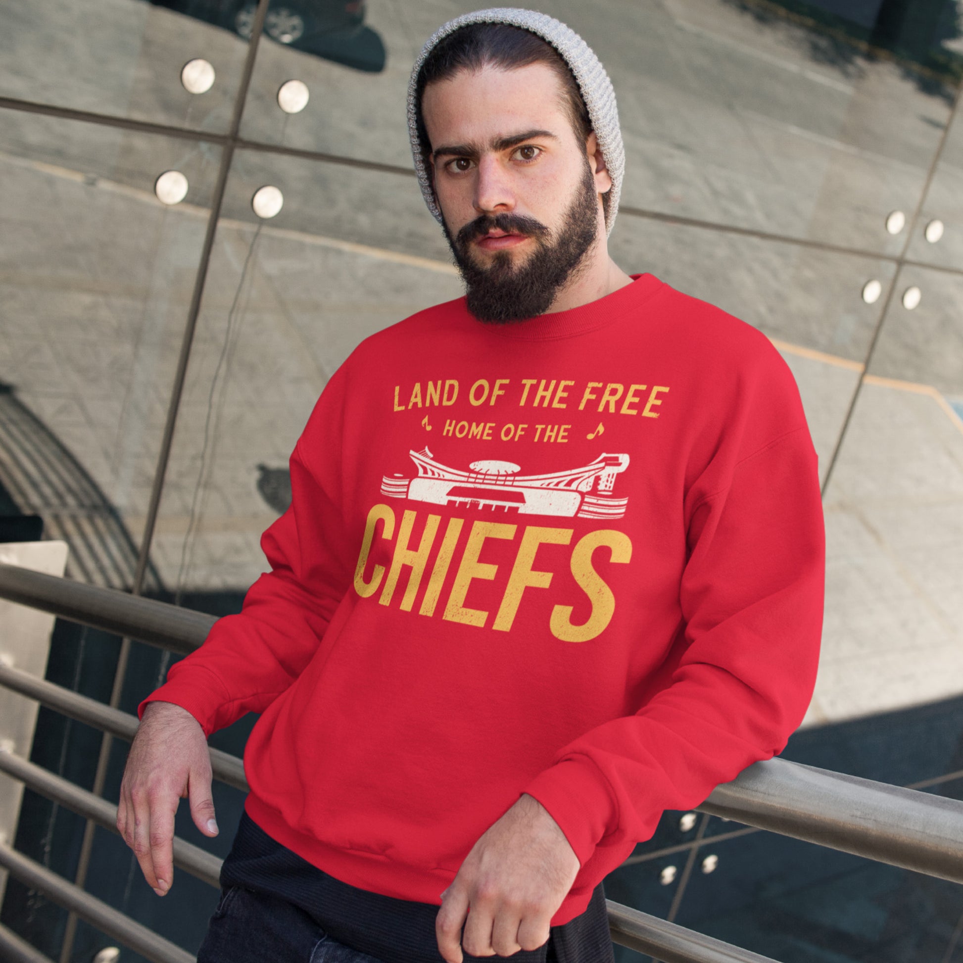 KC Swag Kansas City Chiefs Distressed White & Gold Home Of The Chiefs on a Red Crewneck Sweatshirt worn by a beaning wearing male model leaning on a rail in front of an outdoor mirrored wall