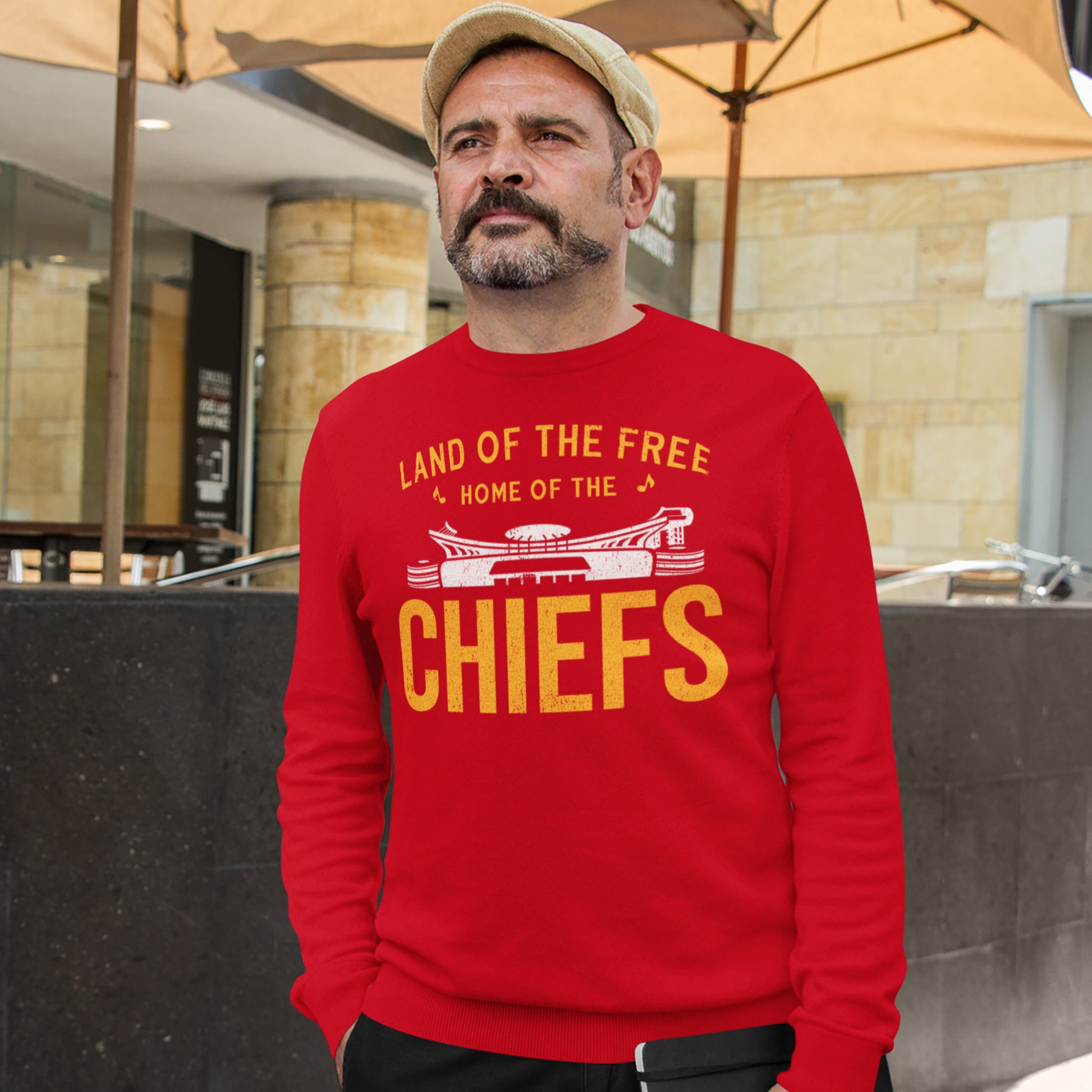 KC Swag Kansas City Chiefs Distressed White & Gold Home Of The Chiefs on a Red Crewneck Sweatshirt worn by a bearded male model wearing a hat walking through an outdoor restaurant patio