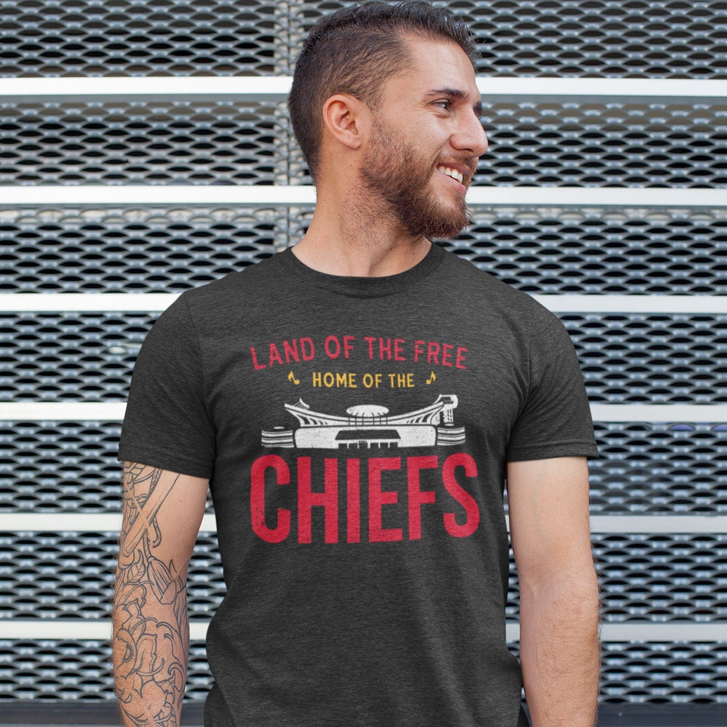 KC Swag | Kansas City Chiefs white & red HOME OF THE CHIEFS on dark heather gray unisex t-shirt worn by male model staning in front of a large metal grate wall