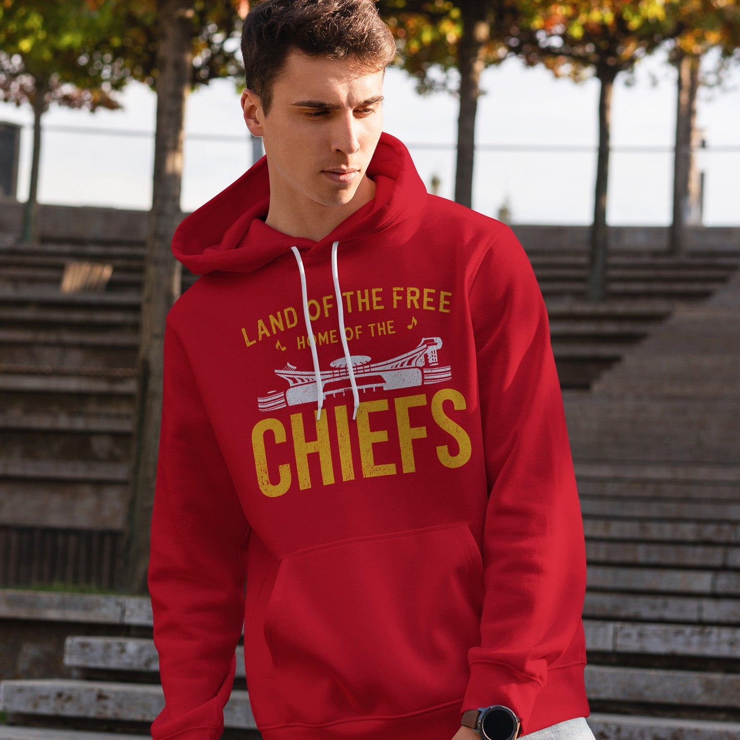 KC Swag | Kansas City Chiefs white/gold HOME OF THE CHIEFS on red sponge-fleece pullover hoodie worn by male model standing on stairs in open-air amphitheater