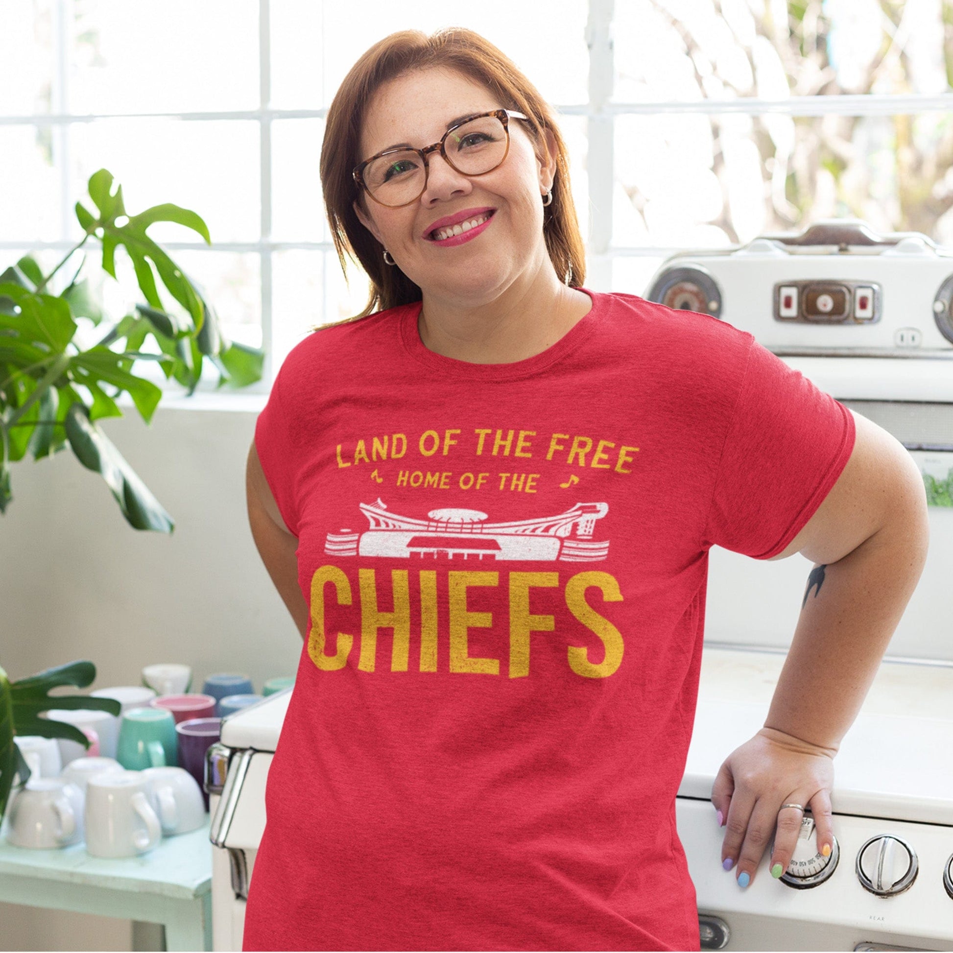 KC Swag | Kansas City Chiefs white & gold HOME OF THE CHIEFS on heather red unisex t-shirt worn by female model leaning against a vintage stove in a bright kitchen