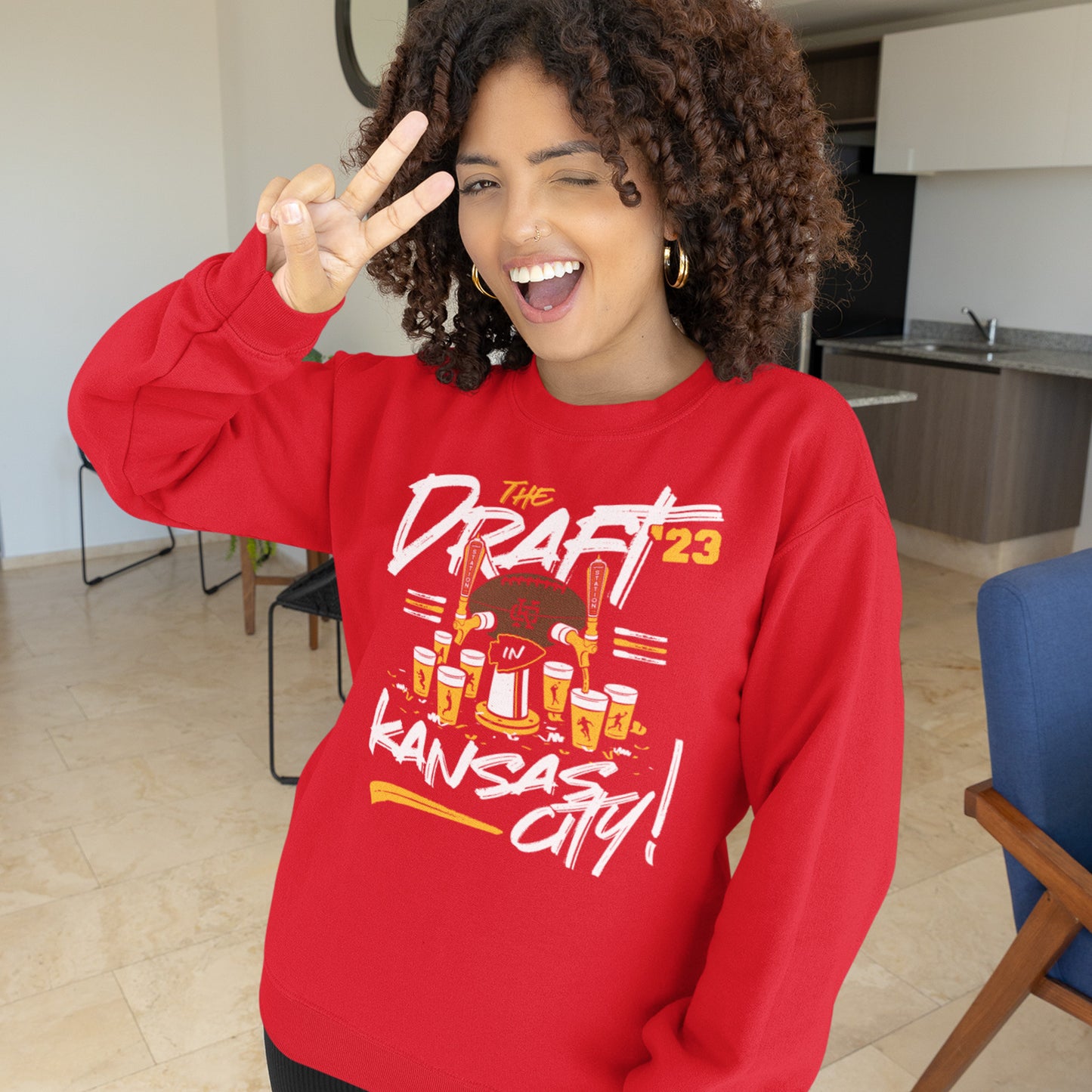 KC Swag Kansas City Chiefs Distressed White & Gold Draftin' IN KC 2023 on a Red Crewneck Sweatshirt worn by curly-haired female model standing in an apartment flashing the peace sign