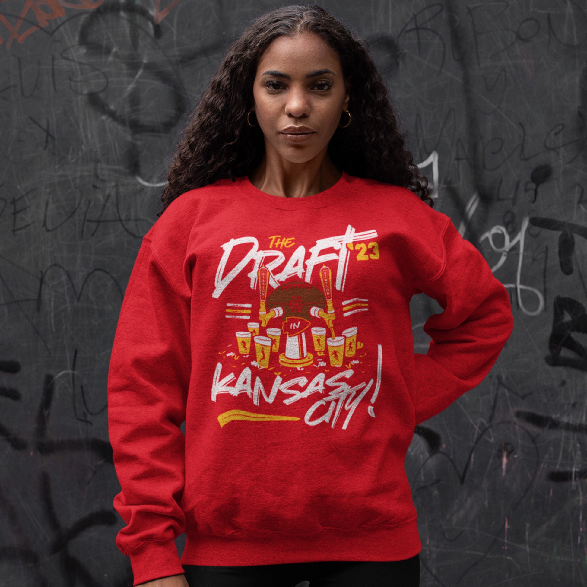 KC Swag Kansas City Chiefs Distressed White & Gold Draftin' IN KC 2023 on a Red Crewneck Sweatshirt worn by a female model standing in front of a dark gray graffitied wall