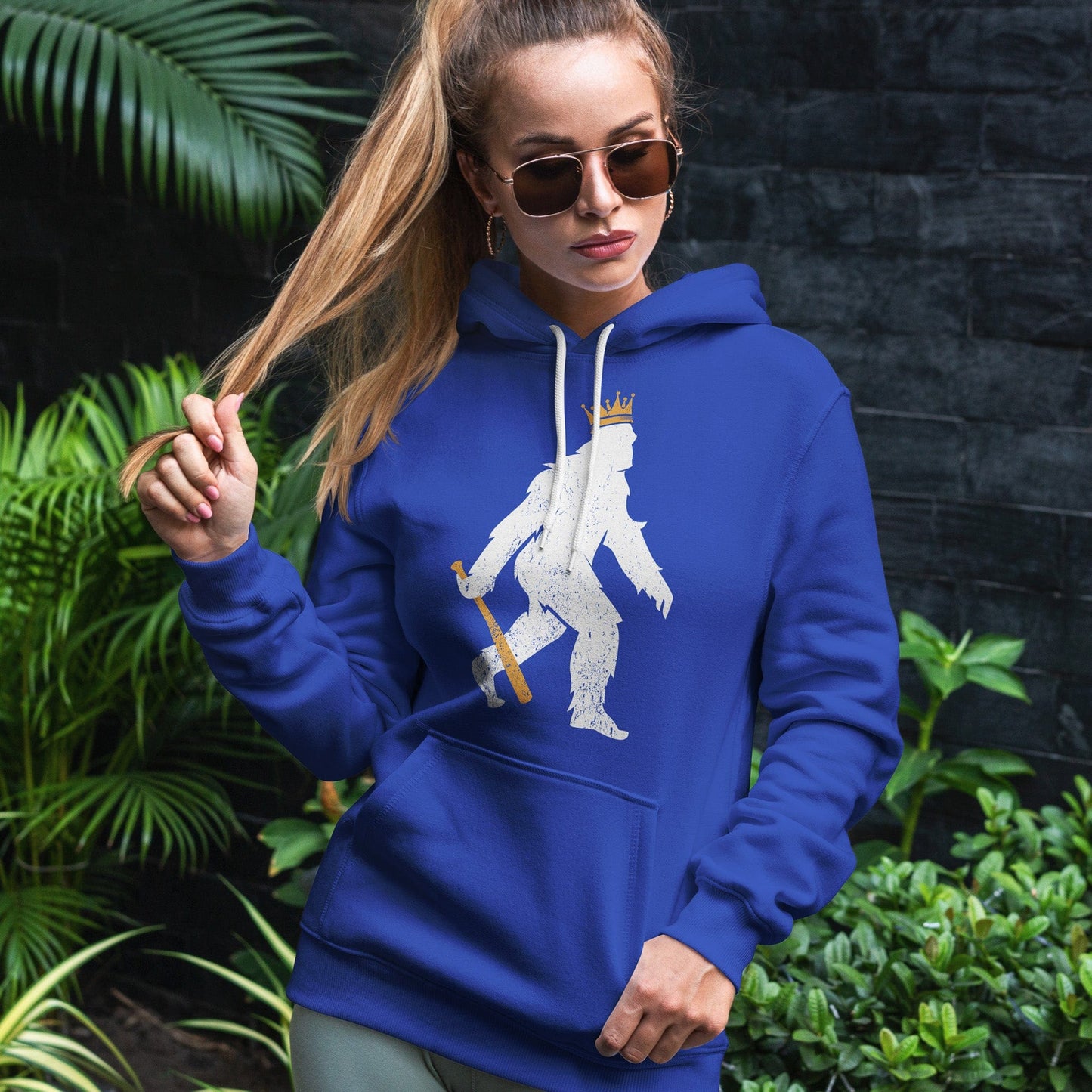 KC Swag | Kansas City Royals white/gold CROWNED PASQUATCH on blue sponge-fleece pullover hoodie worn by female model standing in front of dark gray wall and foliage