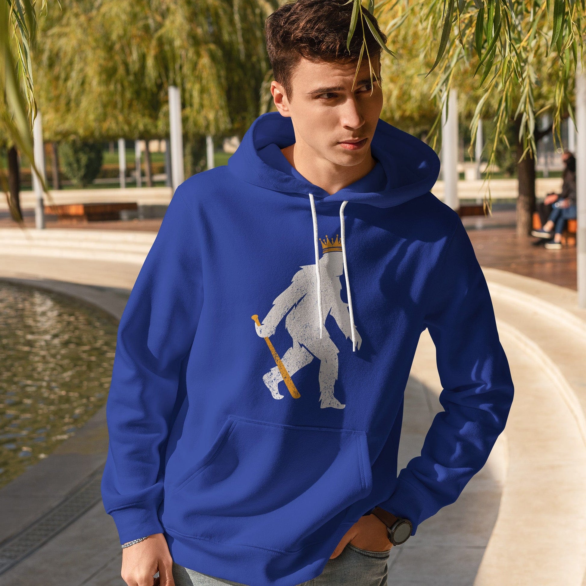 KC Swag | Kansas City Royals white/gold CROWNED PASQUATCH on blue sponge-fleece pullover hoodie worn by male model standing by fountain in a public park
