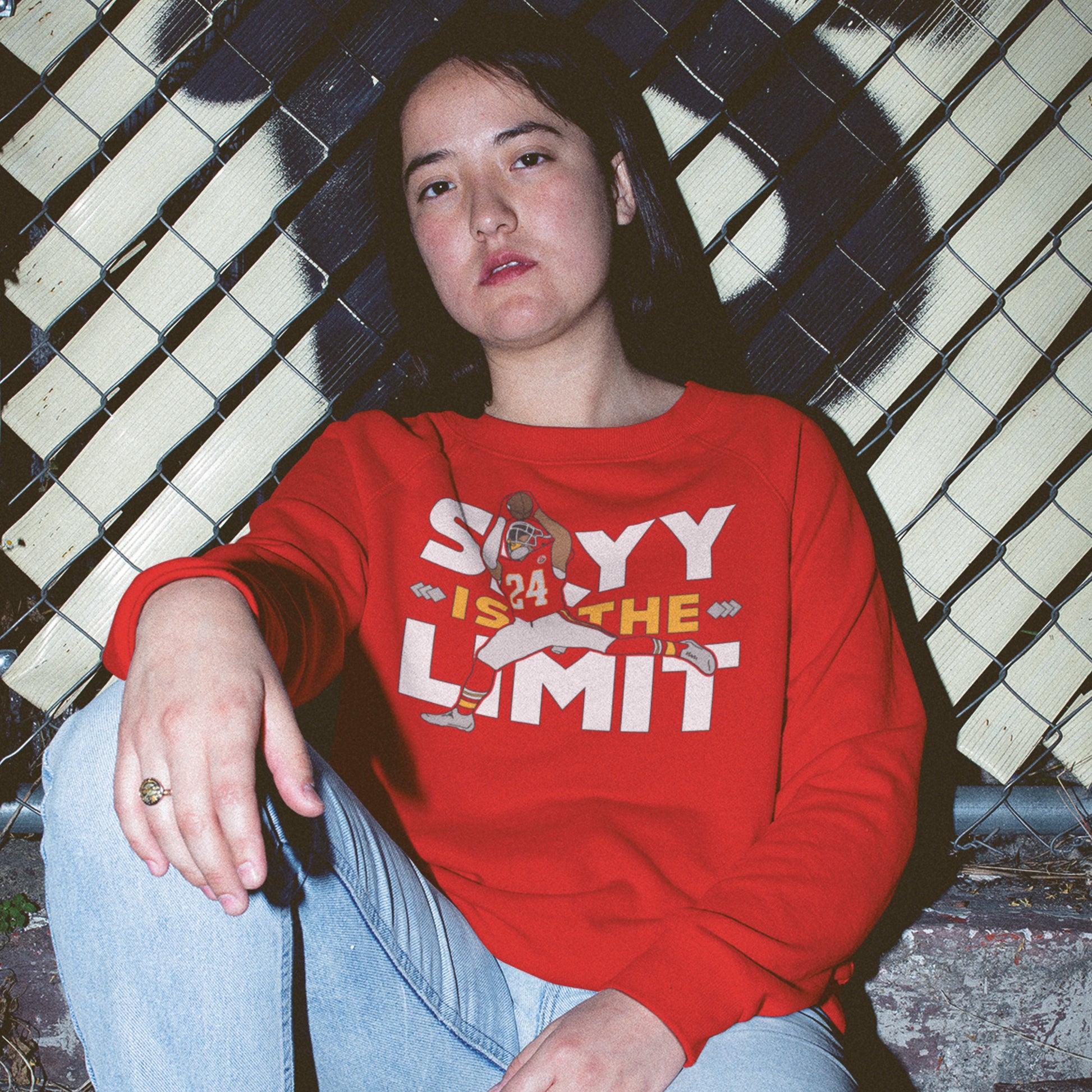 KC Swag Kansas City Chiefs White & Gold Skyy Is The Limit (with football player) on a Red Crewneck Sweatshirt worn by a female model sitting against a chainlink fence in front of a graffitied wall