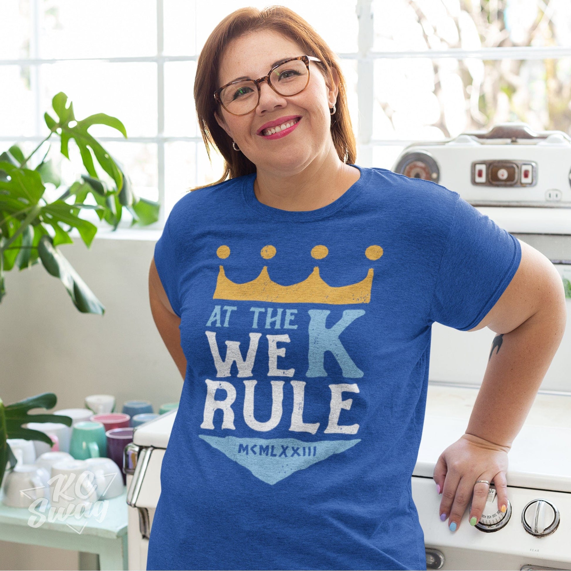 KC Swag Kansas City Royals AT THE K WE RULE on heather royal blue unisex t-shirt worn by female model leaning on stove in bright kitchen