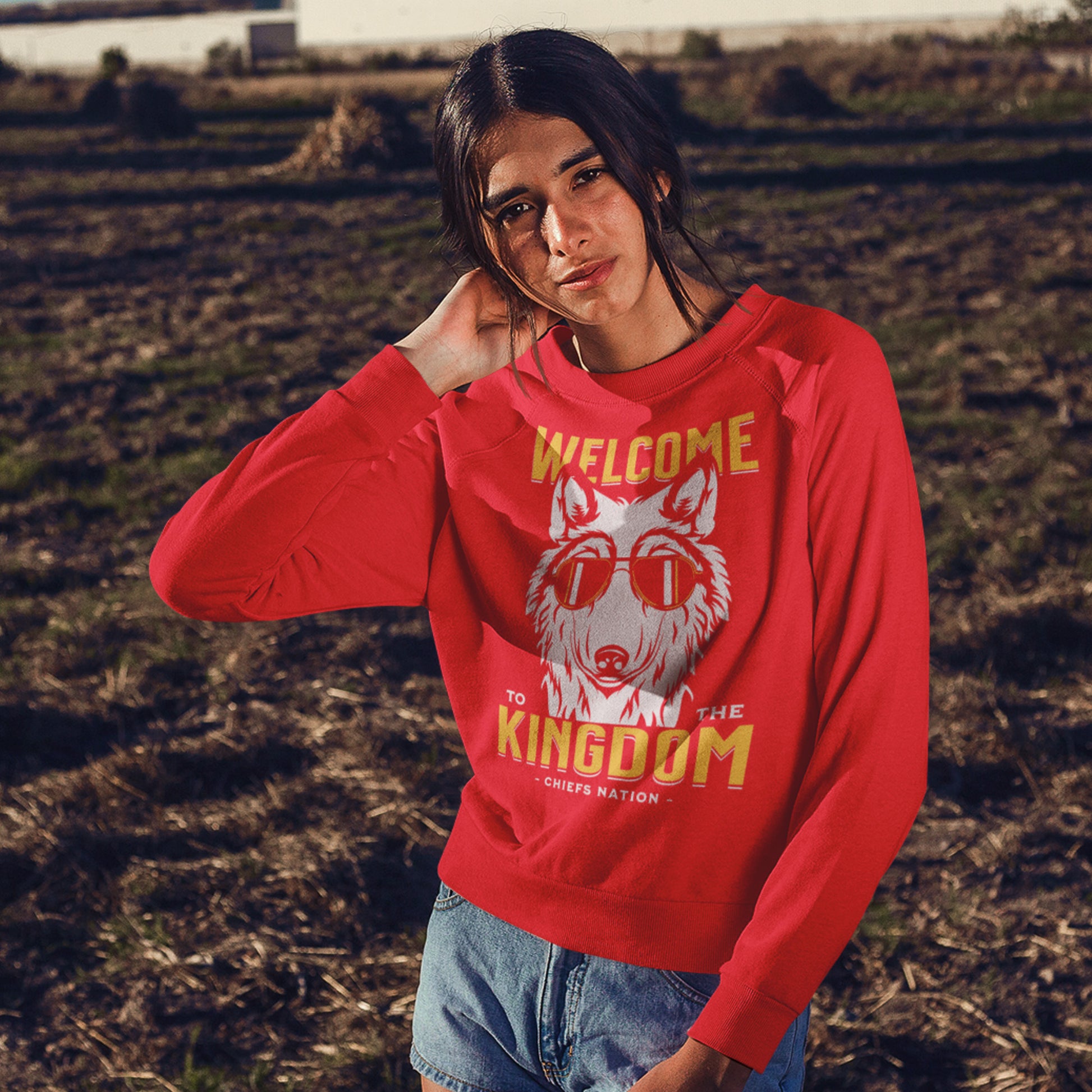 KC Swag Kansas City Chiefs Distressed White & Gold Cool Wolf Kingdom on a Red Crewneck Sweatshirt worn by a dark-haired female m del standing in a barren farmland