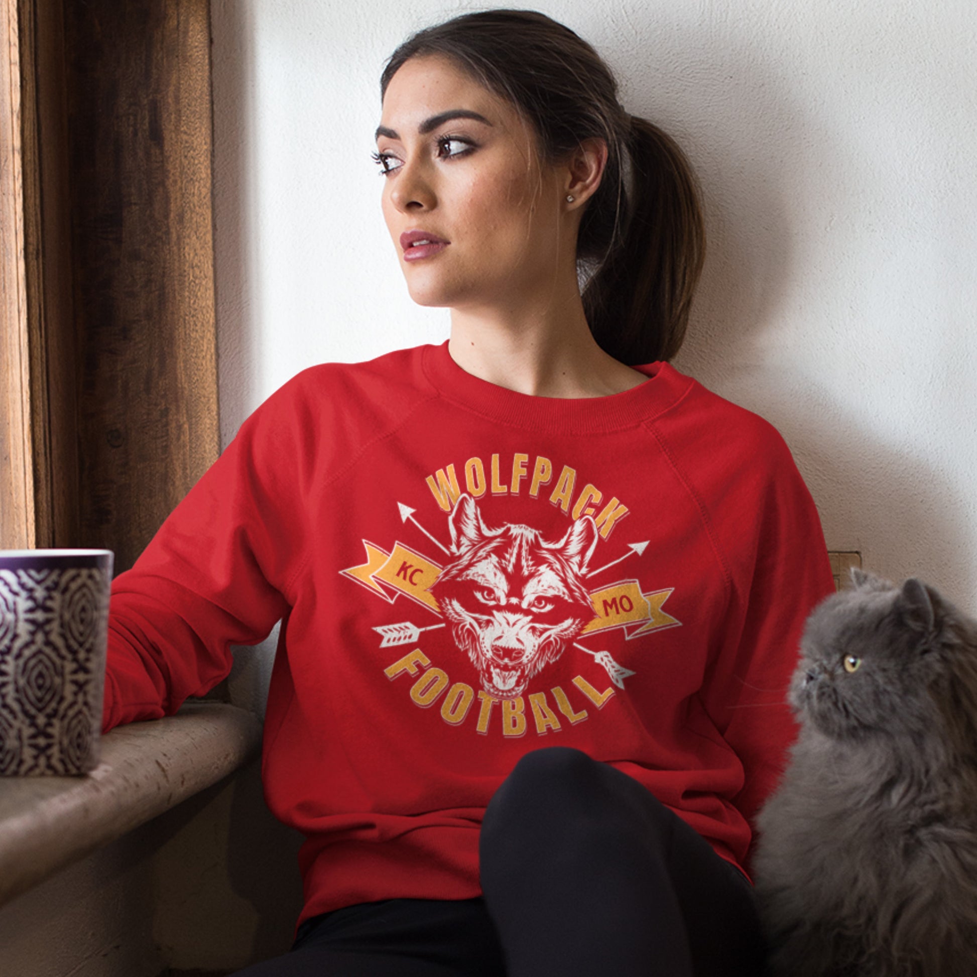 KC Swag Kansas City Chiefs Distressed White & Gold Wolfpack Football on a Red Crewneck Sweatshirt worn by a female model with a mug staring out the window with her cat
