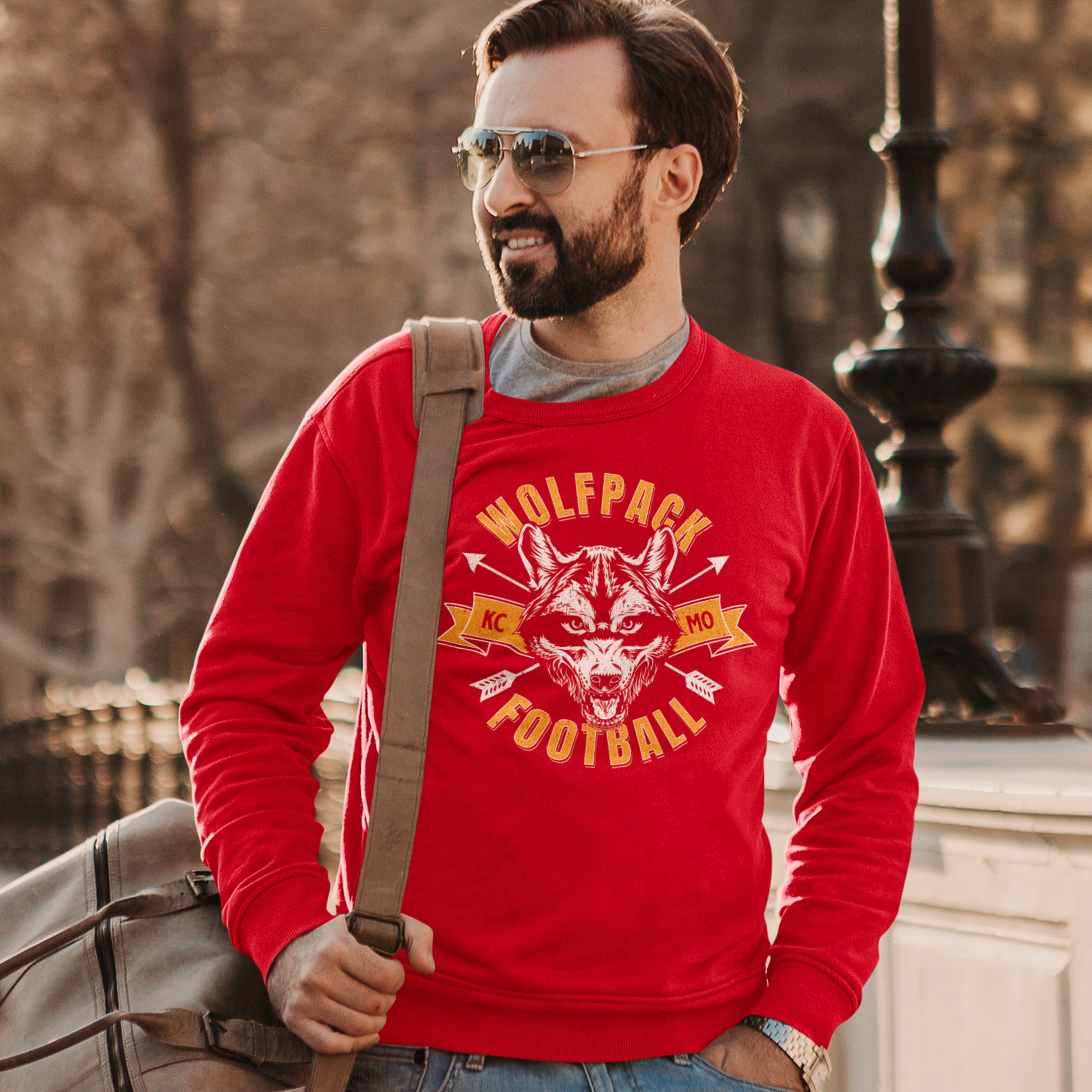 KC Swag Kansas City Chiefs Distressed White & Gold Wolfpack Football on a Red Crewneck Sweatshirt worn by a male model on a bridge carrying a duffle bag over the shoulder