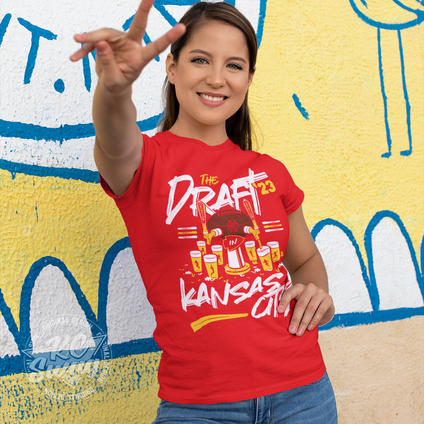 KC Swag - Kansas City Chiefs 2023 NFL DRAFT IN KC UNION STATION on red unisex t-shirt worn by female model standing in front of a graffitied wall flashing the peace sign