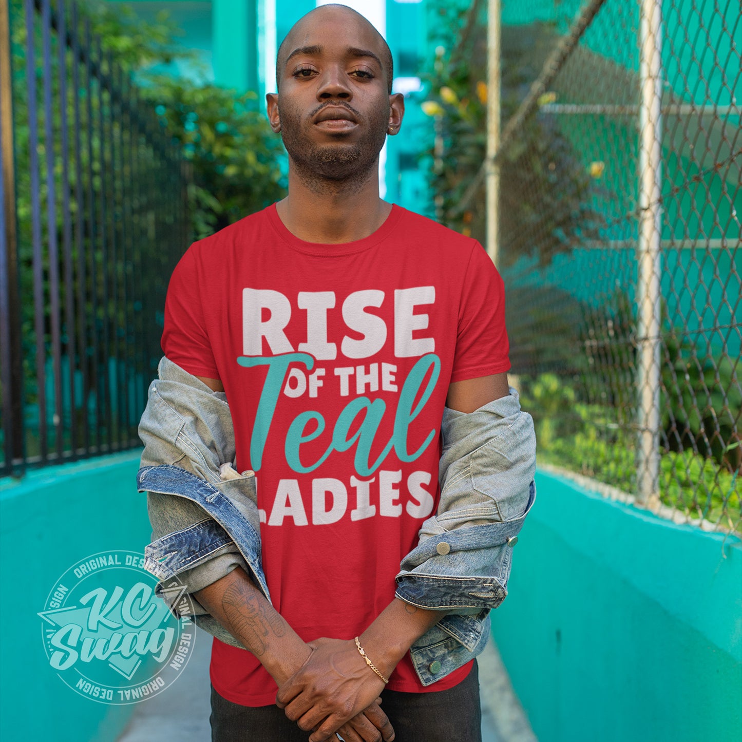 KC Swag - Kansas City Current, Rise Of The Teal Ladies design on a red unisex t-shirt worn by male model standing on a concrete walkway in front of a colorful blue building