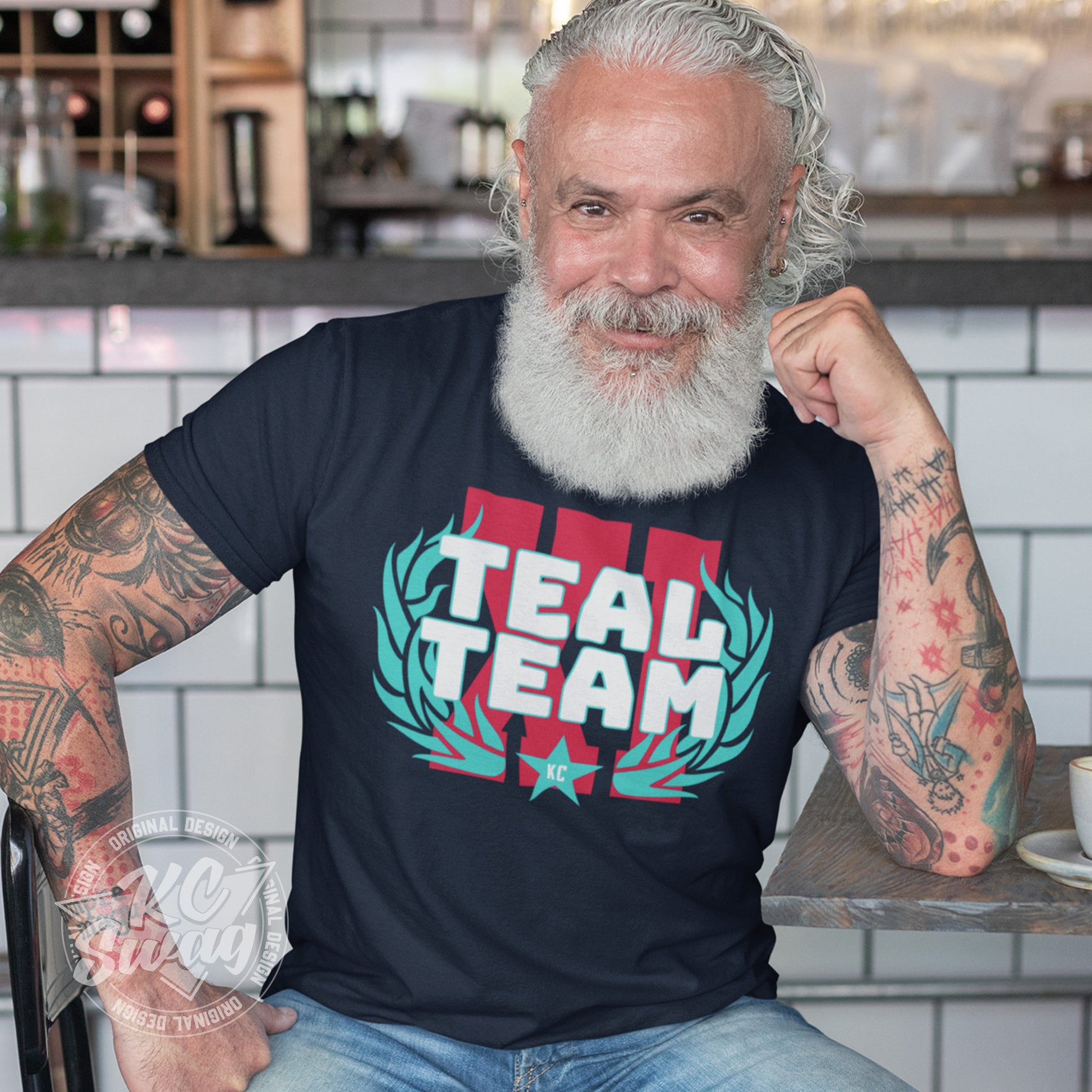 KC Swag - Kansas City Current, Teal Team XI design on navy unisex t-shirt worn by bearded male model sitting at a table in a modern cafe