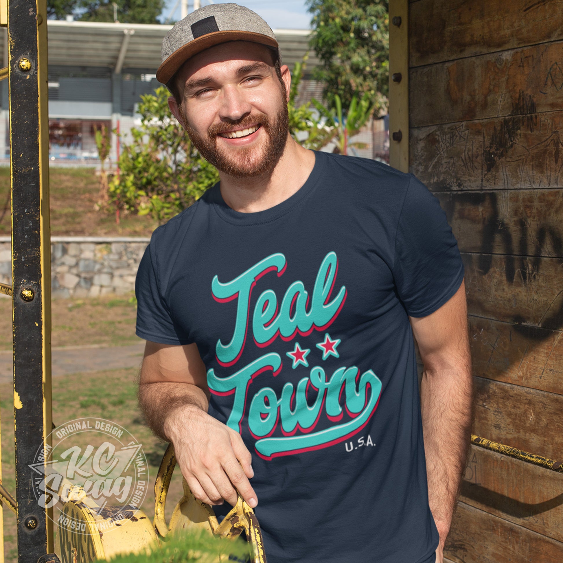 KC Swag - Kansas City Current, Teal Town USA design on navy unisex t-shirt worn by a male model leaning on a rusty garden hose reel wearing a backwards ball cap