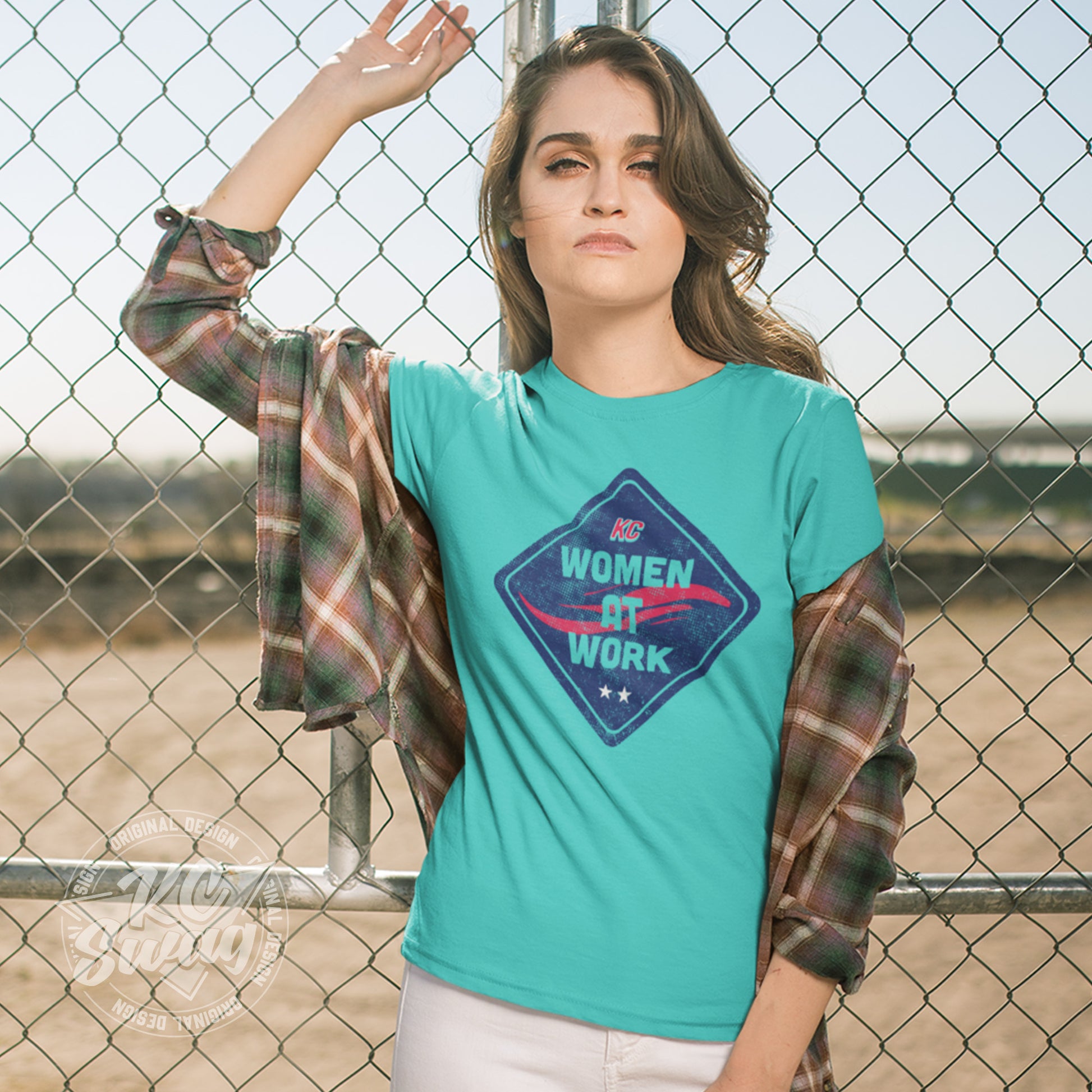 KC Swag - Kansas City Current, Women At Work Construction Sign design on a teal unisex t-shirt worn by female model under open plaid shirt while leaning on a chainlink fence around a construction zone