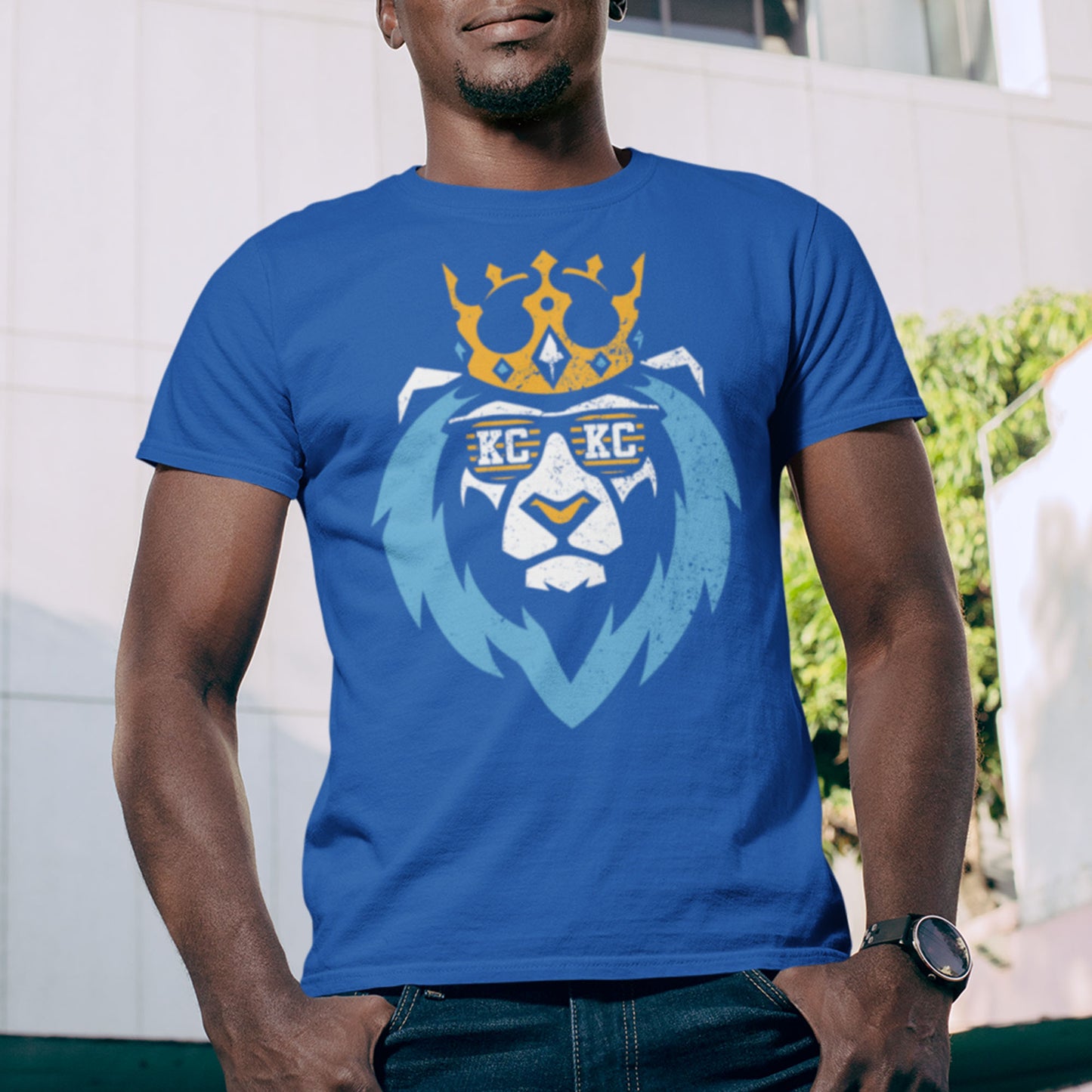 kc swag, kansas city royals, royal blue unisex t-shirt featuring a white and light blue graphic of a lion wearing a gold crown and KC sunglasses, worn by a smiling male model standing on an outdoor platform