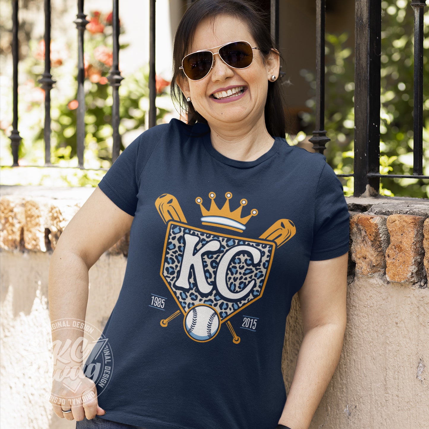 KC Swag - Kansas City Royals, Cheetah Home Plate design on navy blue unisex t-shirt worn by female model leaning on a concrete wall and wearing sunglasses