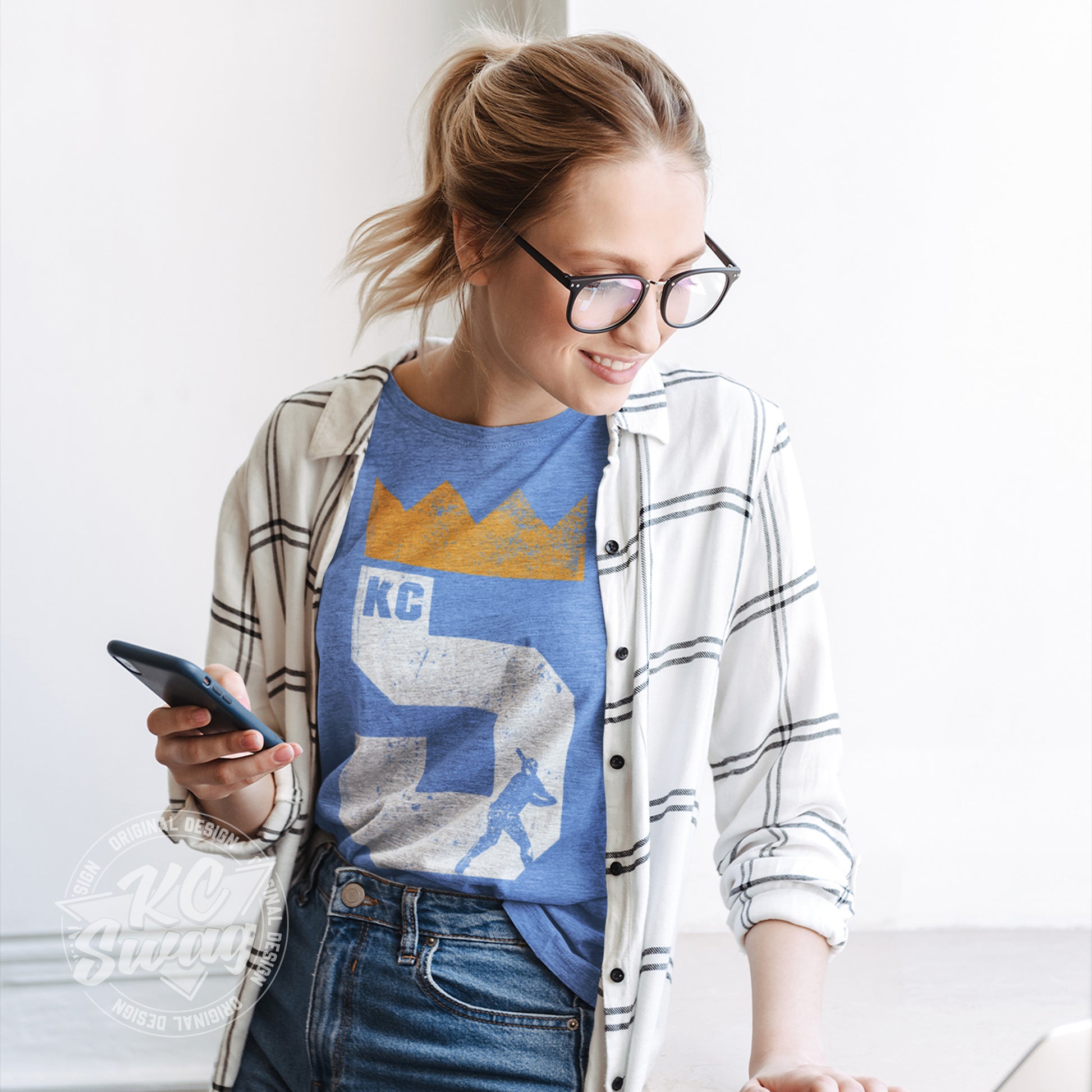 KC Swag - Kansas City Royals, Brett Five Crown design on heather columbia blue unisex t-shirt worn by female model under an open plaid shirt while looking away from her phone in an office setting