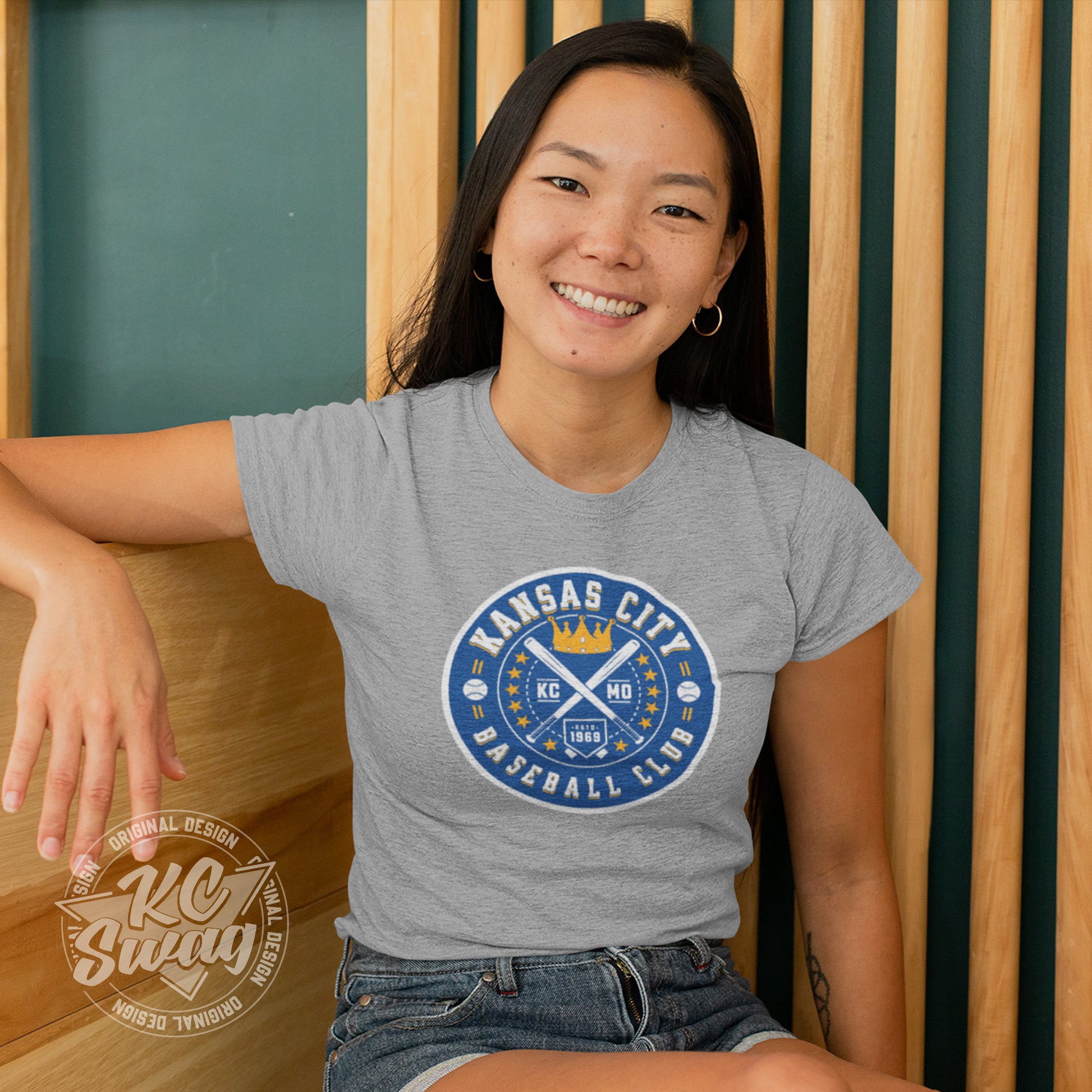 KC Swag - Kansas City Royals, Baseball Club design on athletic heather grey unisex t-shirt worn by female model sitting on wood bench in front of a wood slatted wall