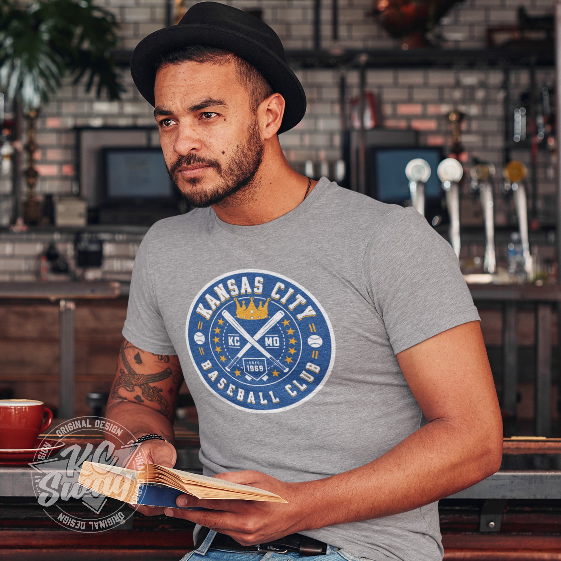 KC Swag - Kansas City Royals, Baseball Club design on athletic heather grey unisex t-shirt worn by male model sitting at the bar in an retro cafe
