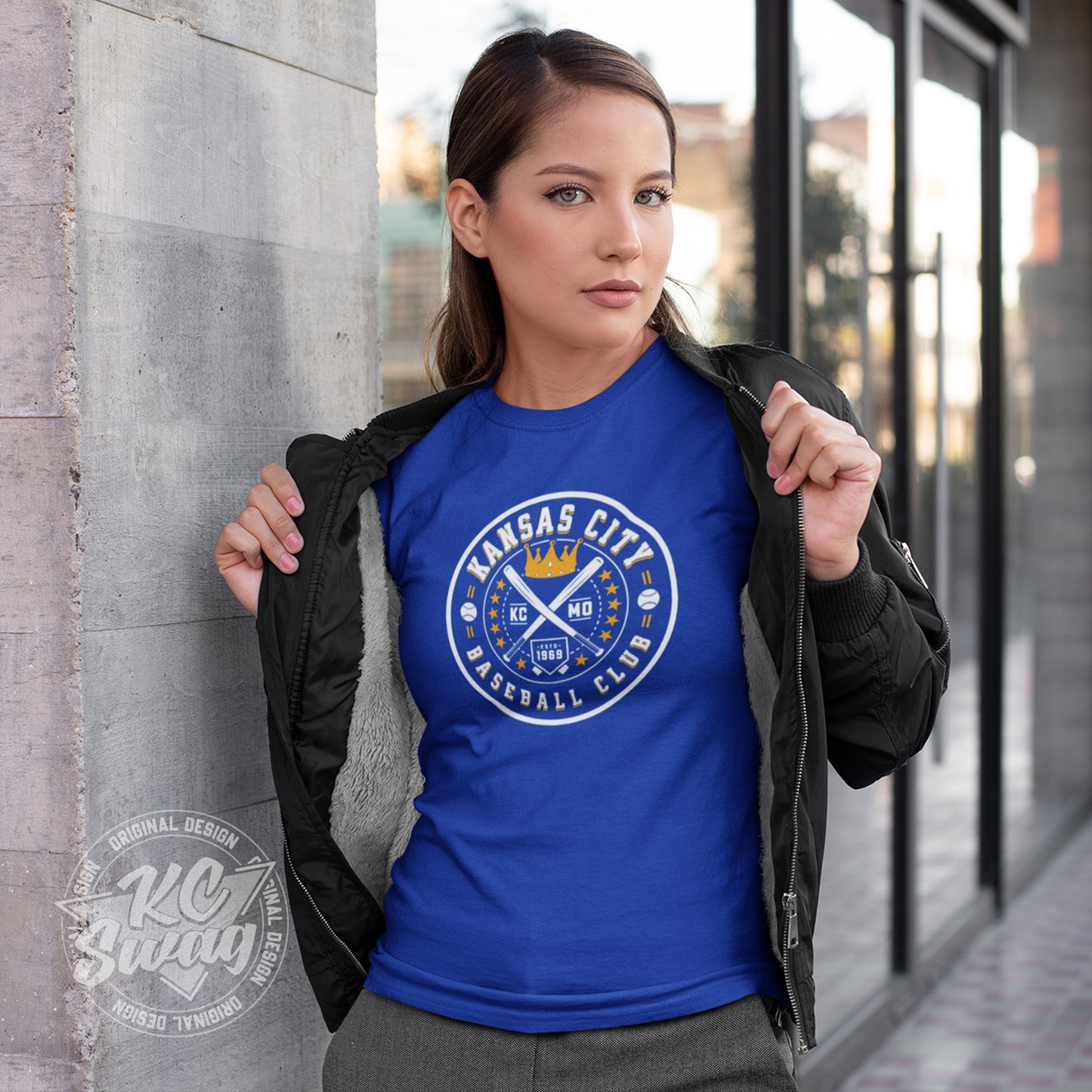 KC Swag - Kansas City Royals, Baseball Club design on royal blue unisex t-shirt worn by female model under open jacket leaning on a concrete pillar