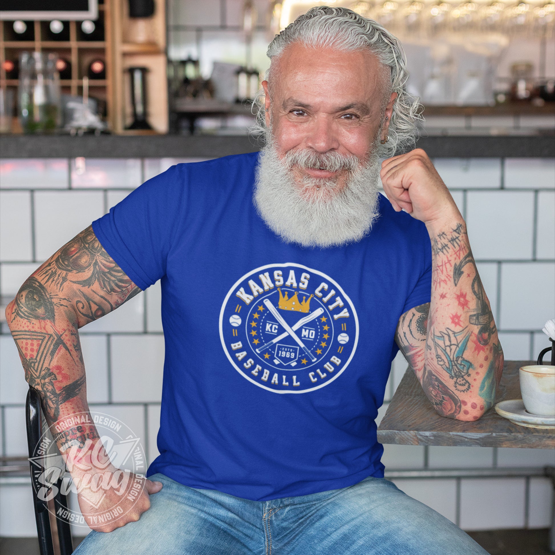 KC Swag - Kansas City Royals, Baseball Club design on royal blue unisex t-shirt worn by bearded male model sitting at a table in a modern cafe