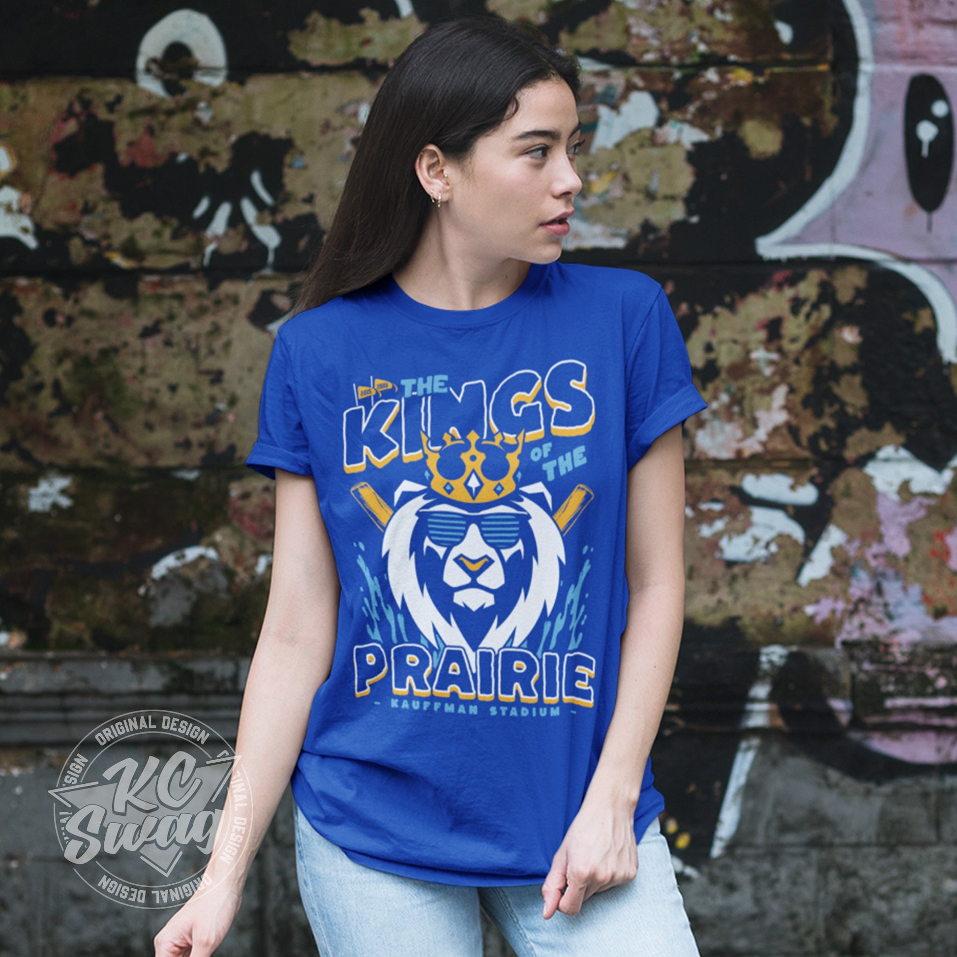 KC Swag - Kansas City Royals, Kings Of The Prairie design on royal blue unisex t-shirt worn by female model standing in front of a grunge graffitied wall in an urban alley