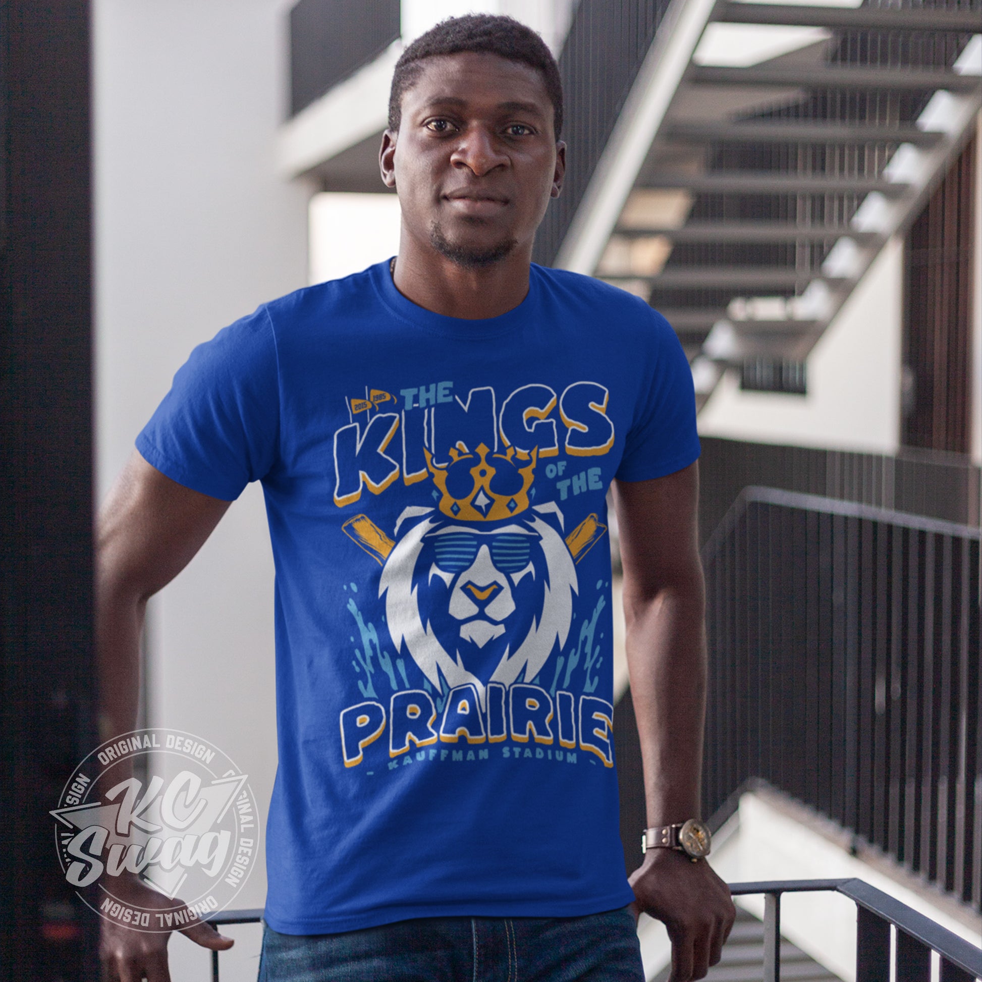 KC Swag - Kansas City Royals, Kings Of The Prairie design on royal blue unisex t-shirt worn by male model leaning on railing on an apartment balcony