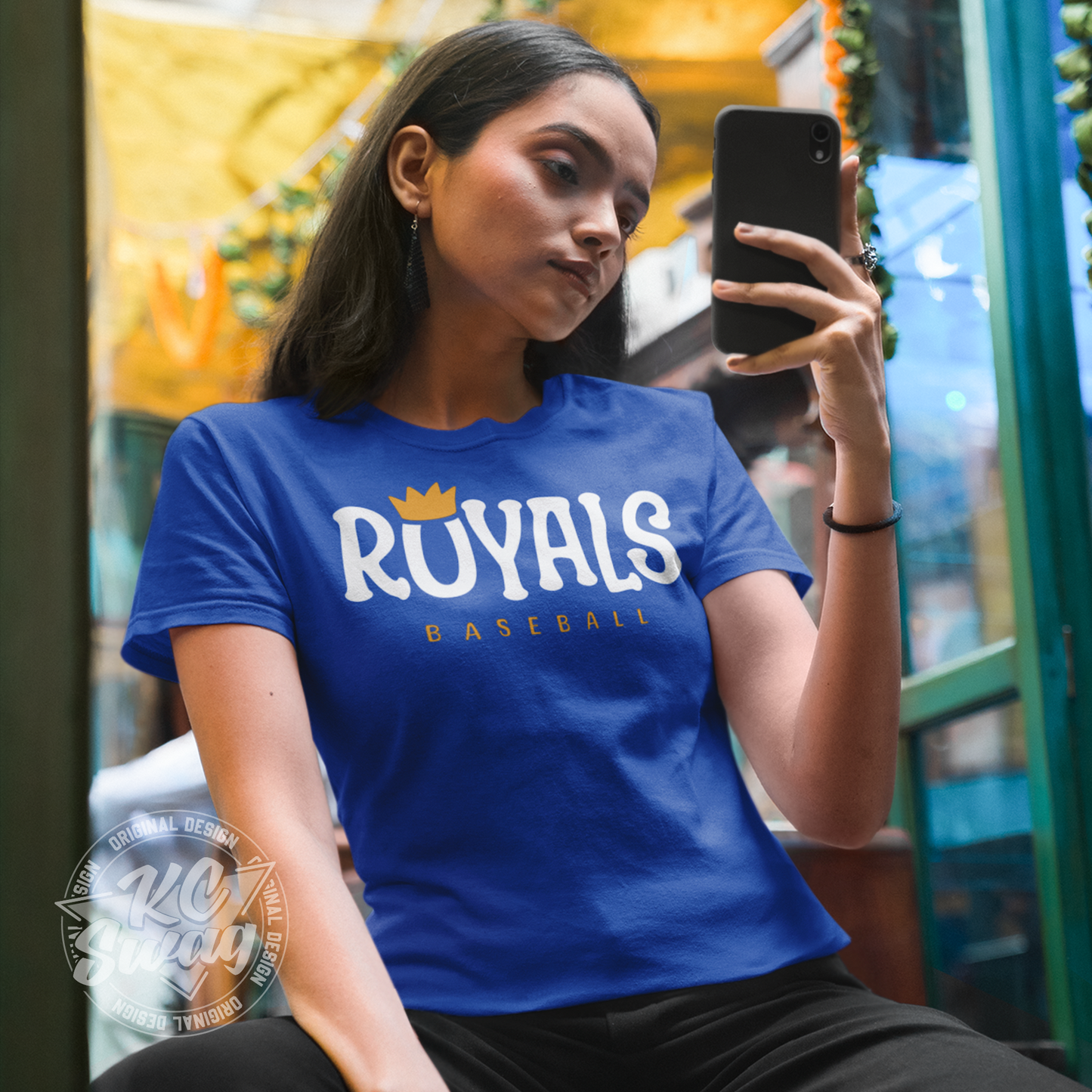 KC Swag - Kansas City Royals, Royals O-Crown design on royal blue unisex t-shirt worn by female model taking a mirror selfie in front of color draped fabrics