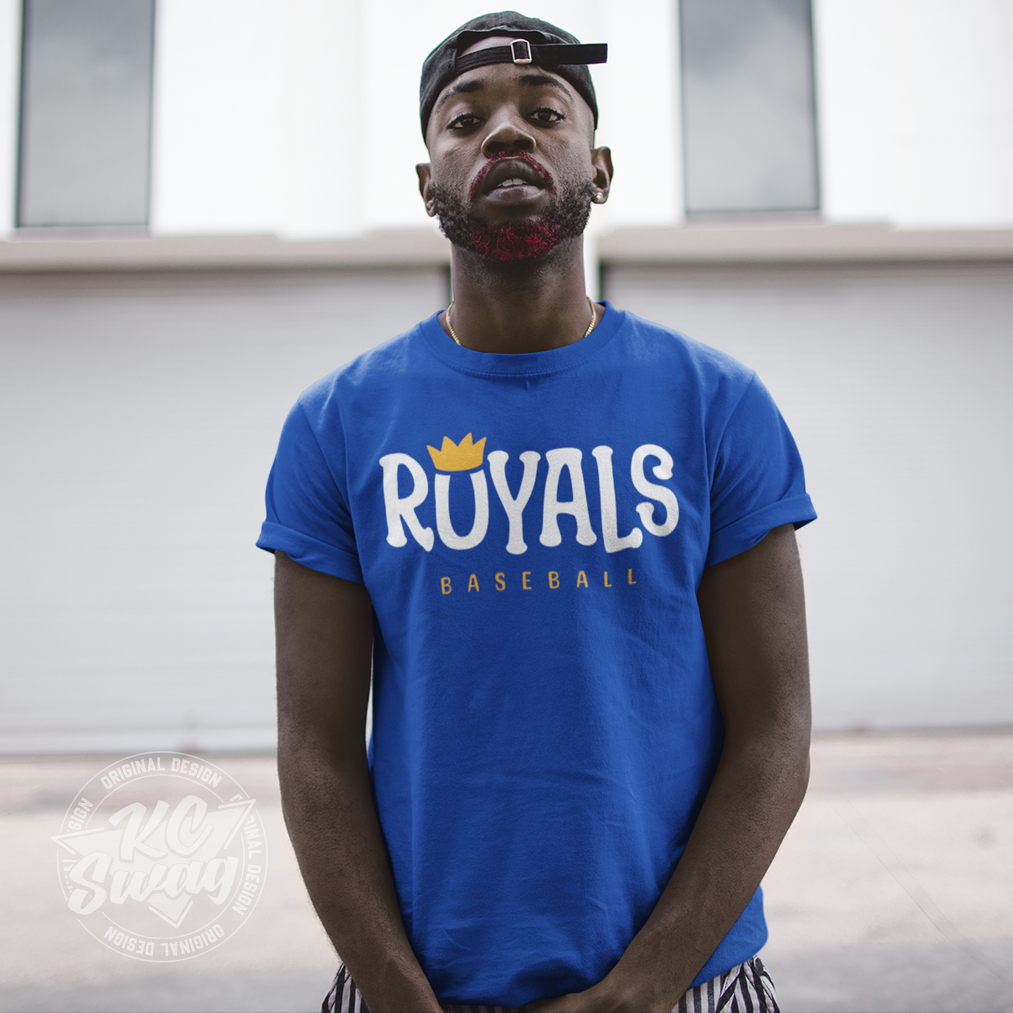 KC Swag - Kansas City Royals, Royals O-Crown design on royal blue unisex t-shirt worn by male model standing in an urban alley with a backwards baseball cap