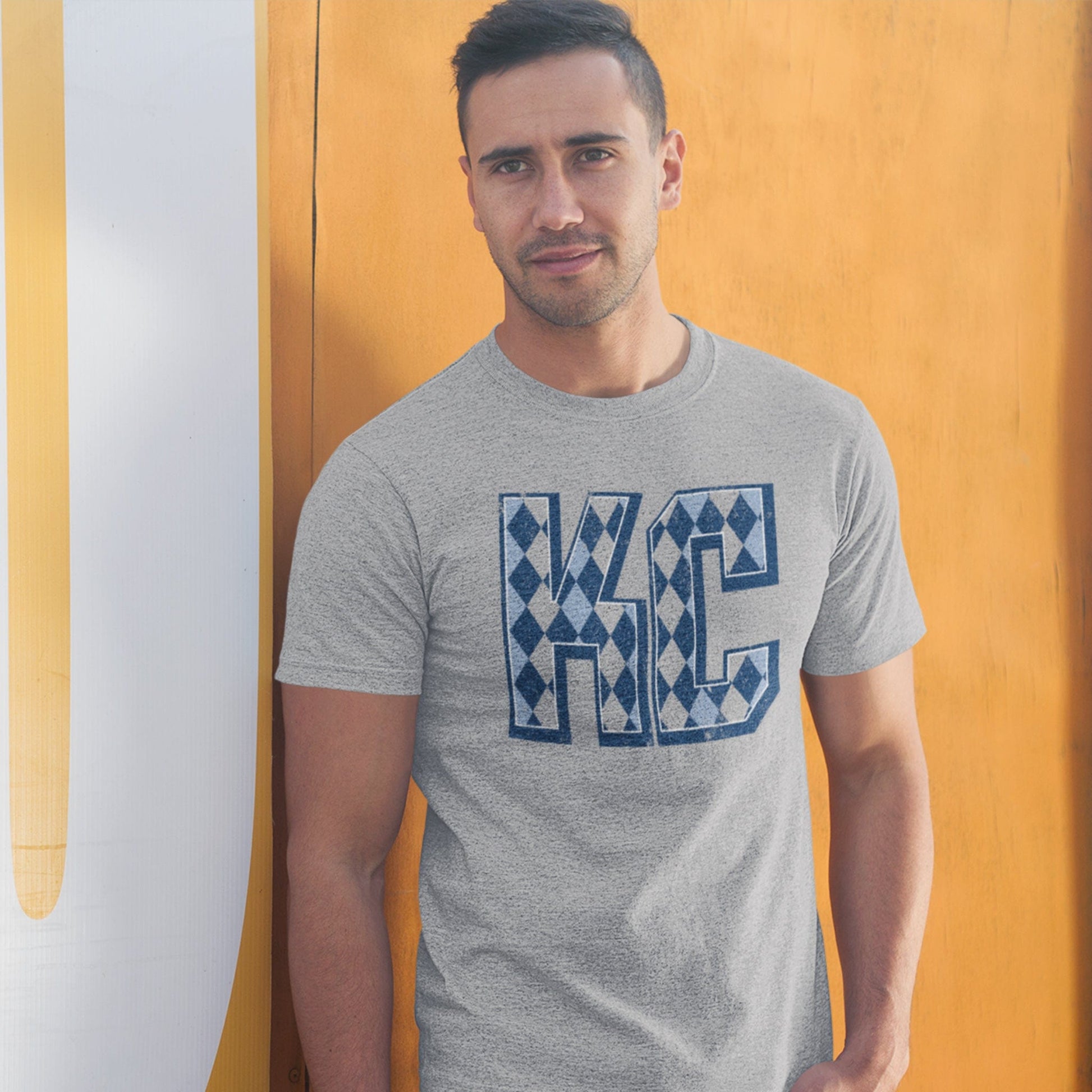 KC Swag Sporting Kansas City powder blue/navy/grey ARGYLE PATTERN KC on athletic heather grey t-shirt worn by male model leaning against yellow wall