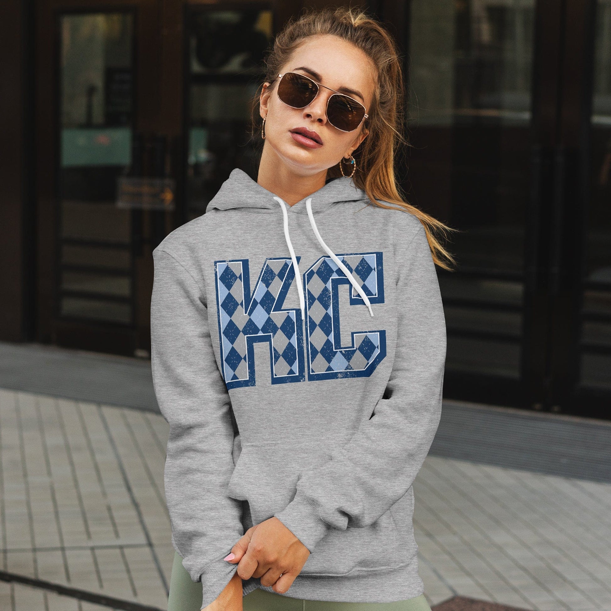 KC Swag Sporting Kansas City powder blue/navy/grey/white ARGYLE pattern KC on athletic heather grey fleece pull-over hoodie worn by female model outside of department store doors