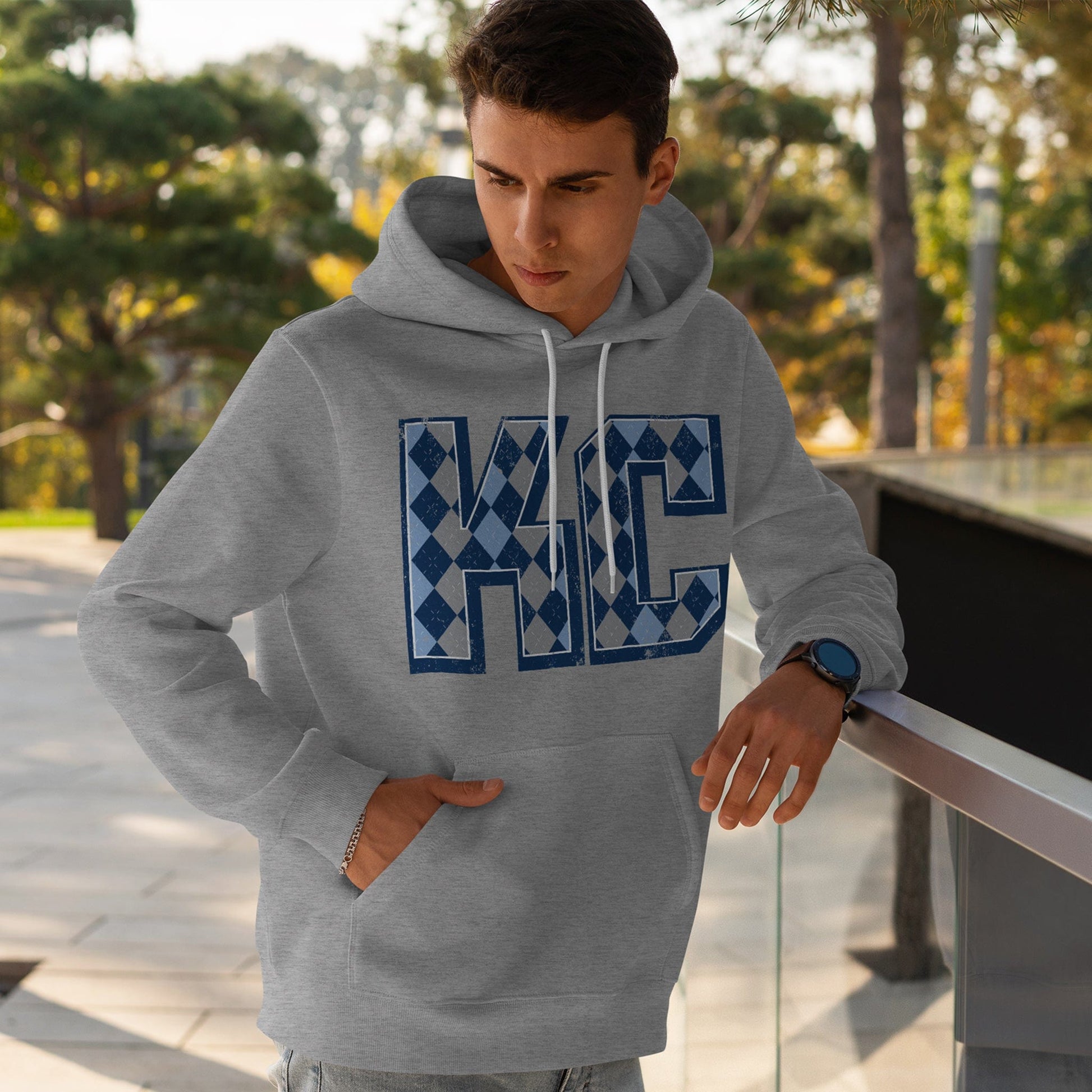 KC Swag Sporting Kansas City powder blue/navy/grey/white ARGYLE pattern KC on athletic heather grey fleece pull-over hoodie worn by male model leaning on rail in outdoor plaza