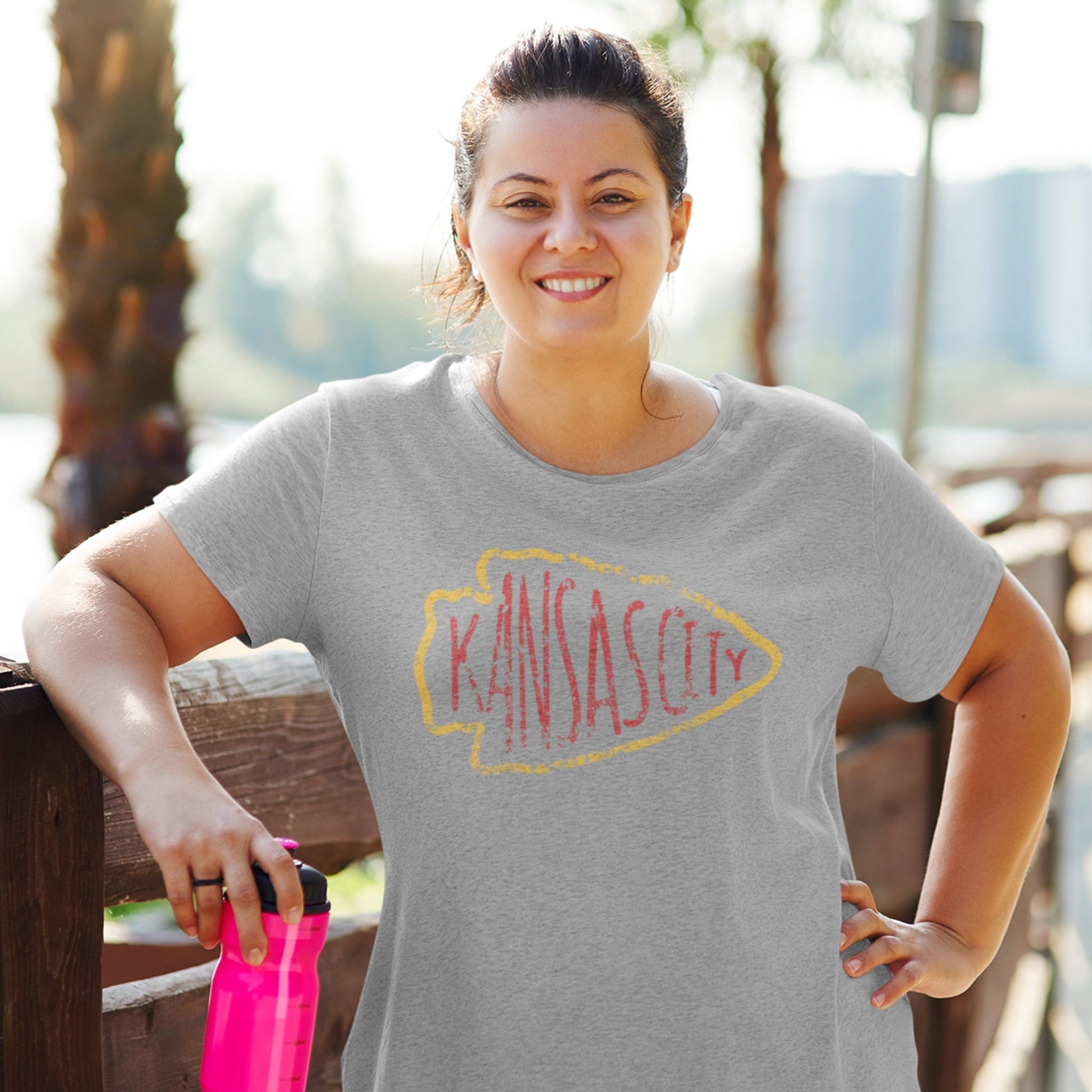 KC Swag Kansas City Chiefs red KANSA CITY inside yellow arrowhead outline on athletic heather grey t-shirt worn by female model in sunny outdoor setting