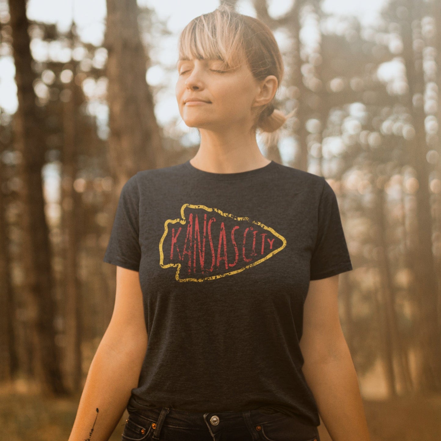 KC Swag Kansas City Chiefs red KANSA CITY inside yellow arrowhead outline on dark heather grey t-shirt worn by female model in warm forest setting
