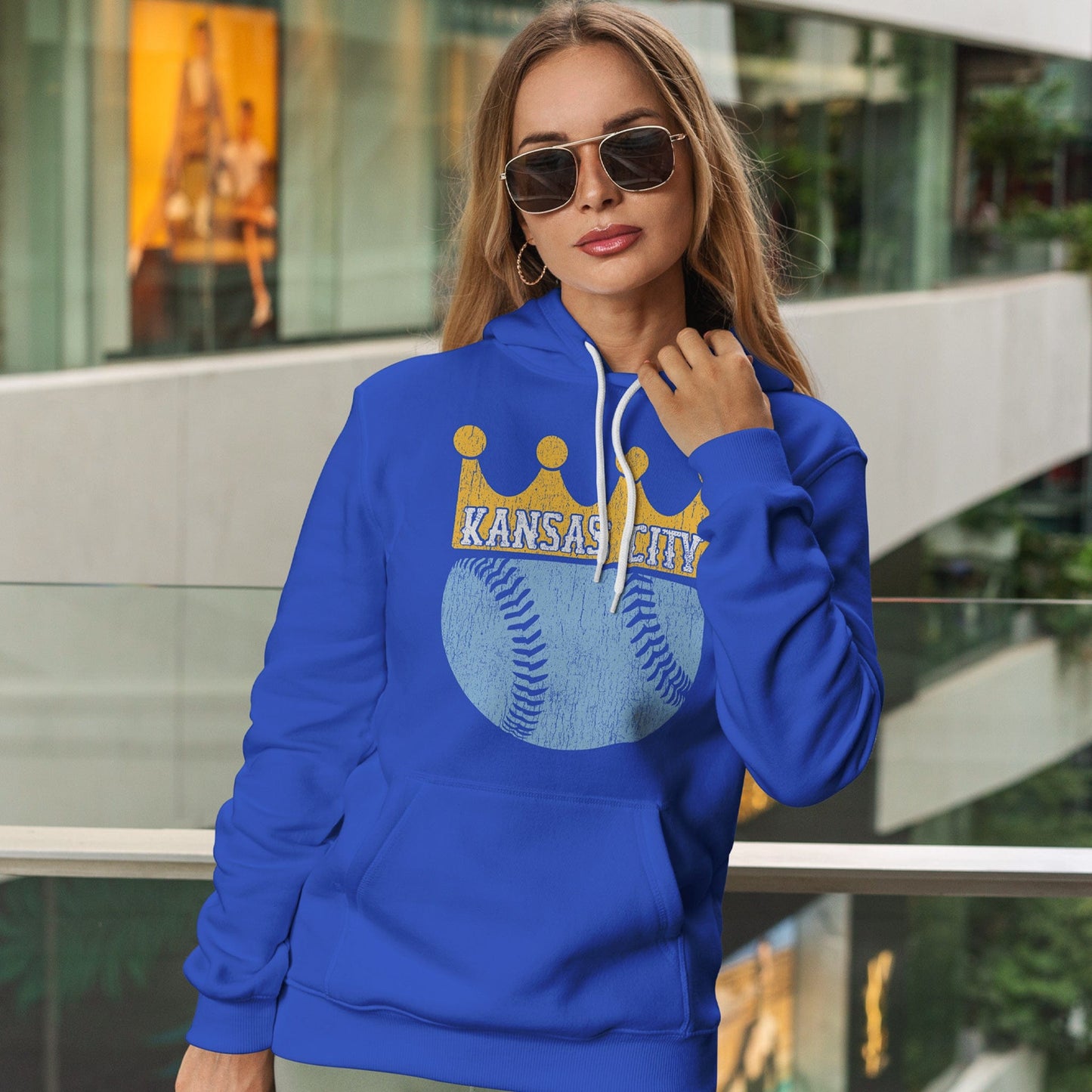 KC Swag Kansas City Royals powder blue baseball wearing a gold crown with KANSAS CITY text on royal blue pull-over hoodie worn by female model inside shopping mall