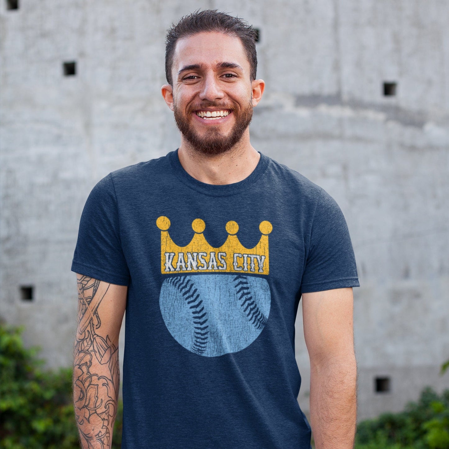 KC Swag Kansas City Royals distressed gold/light-blue BASEBALL CROWN that reads KANSAS CITY in white on a soft heather navy t-shirt worn by male model in front of concrete wall