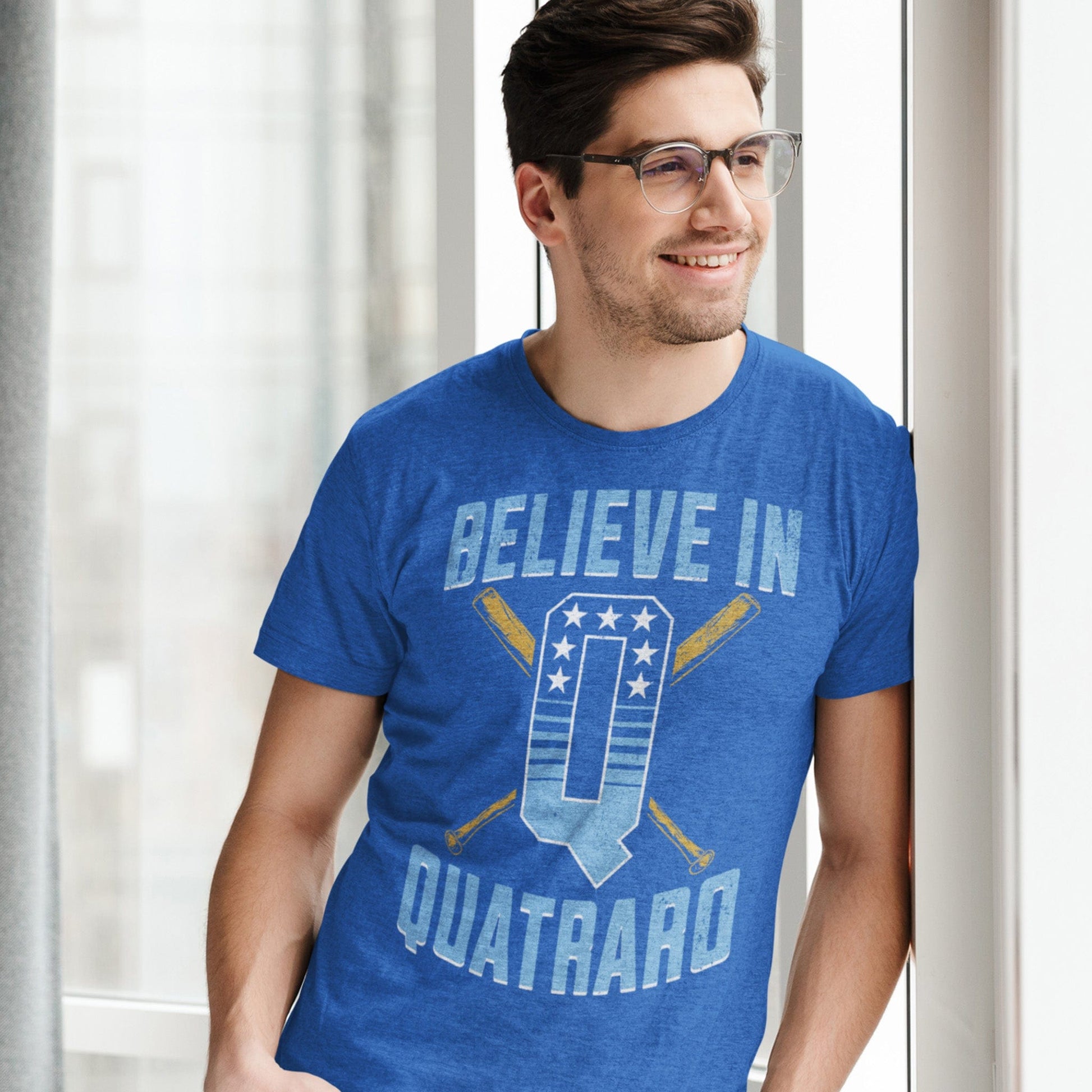 KC Swag Kansas City Royals royal blue, powder, gold, white BELIEVE IN Q on heather royal blue unisex t-shirt worn by male model leaning on wall near window in apartment