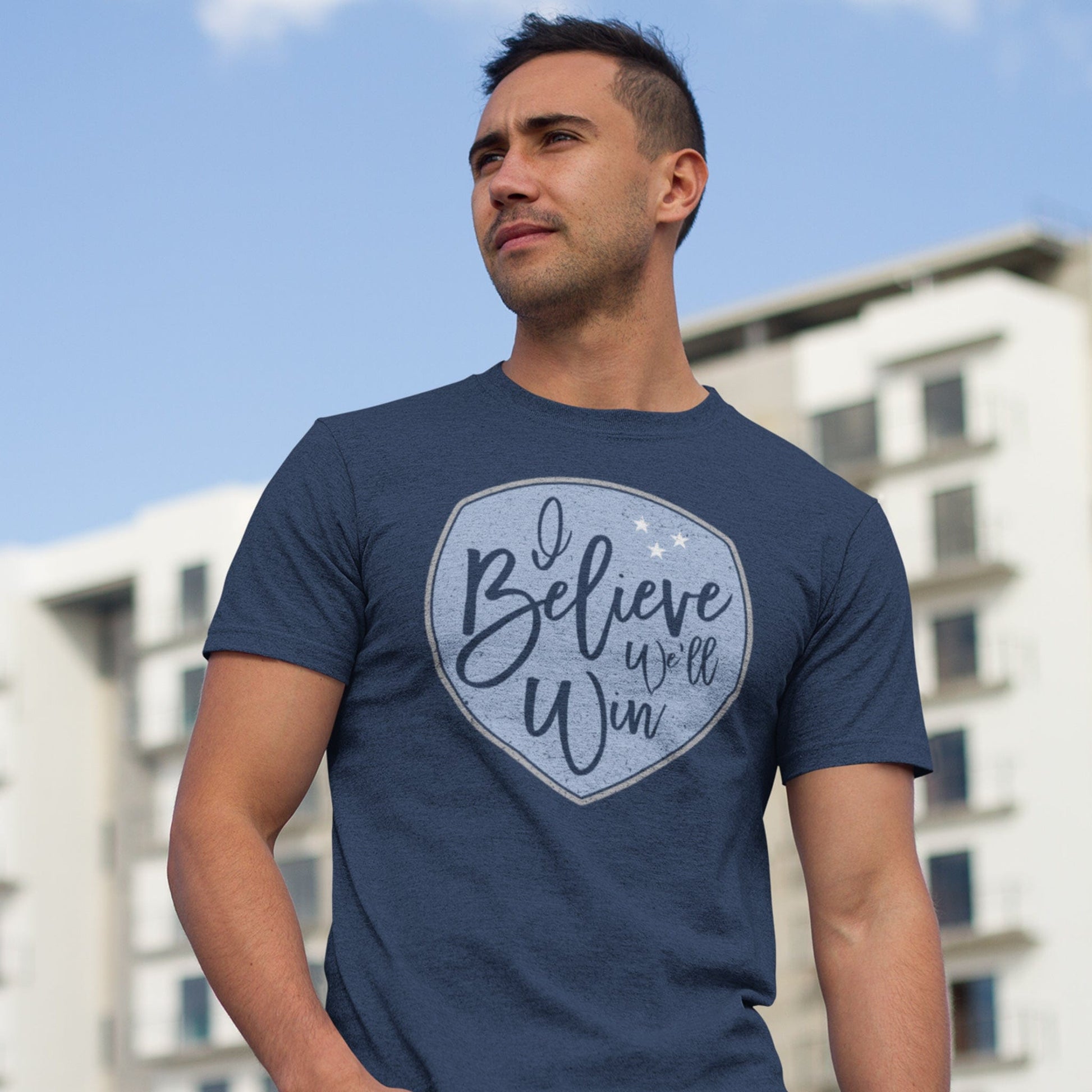 KC Swag Sporting Kansas City light blue/grey/white I BELIEVE WE'LL WIN shield on heather navy t-shirt worn by male model in front of apartment building
