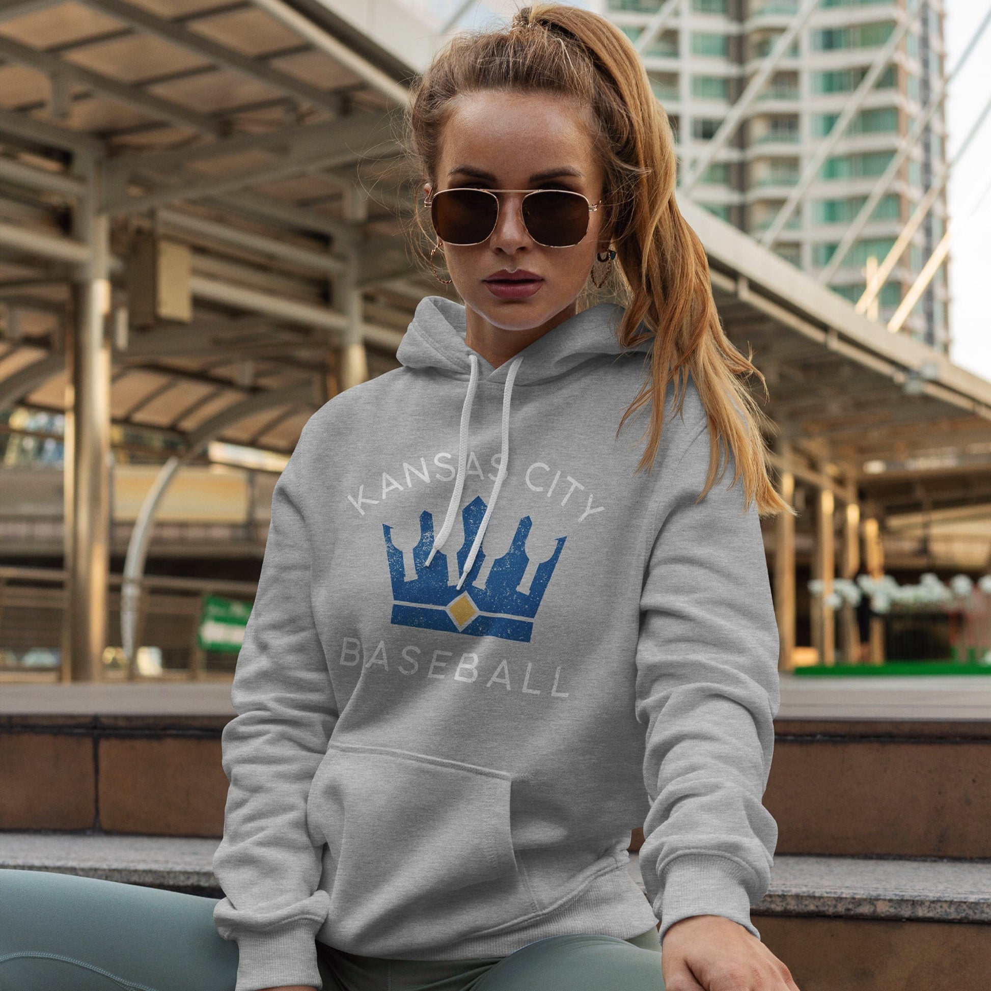KC Swag Kansas City Royalsroyal-blue/gold BOTTLE CROWN with white KC ROYALS text on athletic heather grey pull-over hoodie worn by female model sitting in front of downtown buildings