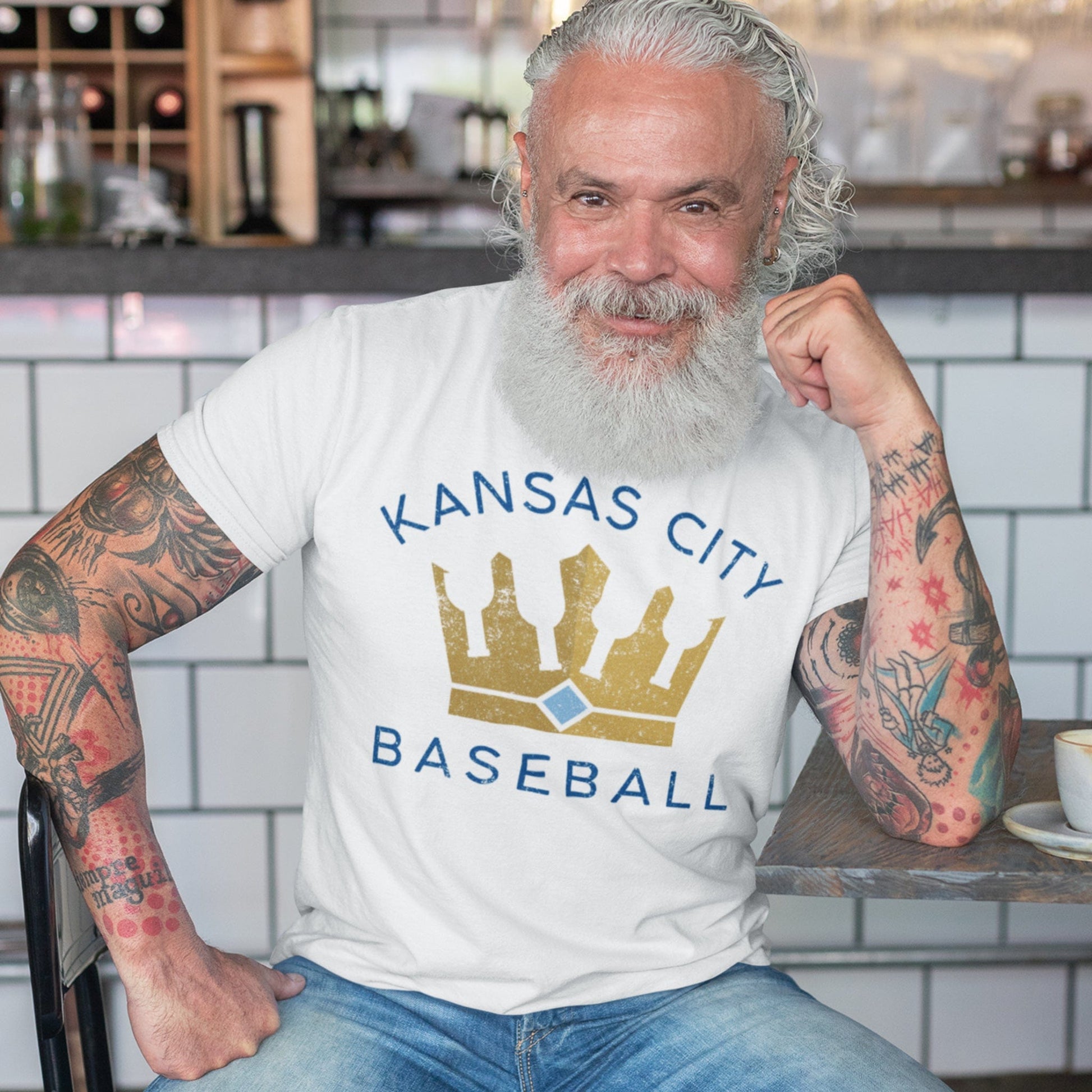 KC Swag Kansas City Royals gold/blue KANSAS CITY BASEBALL BOTTLE CROWN on white t-shirt worn by grey bearded male model sitting in cafe