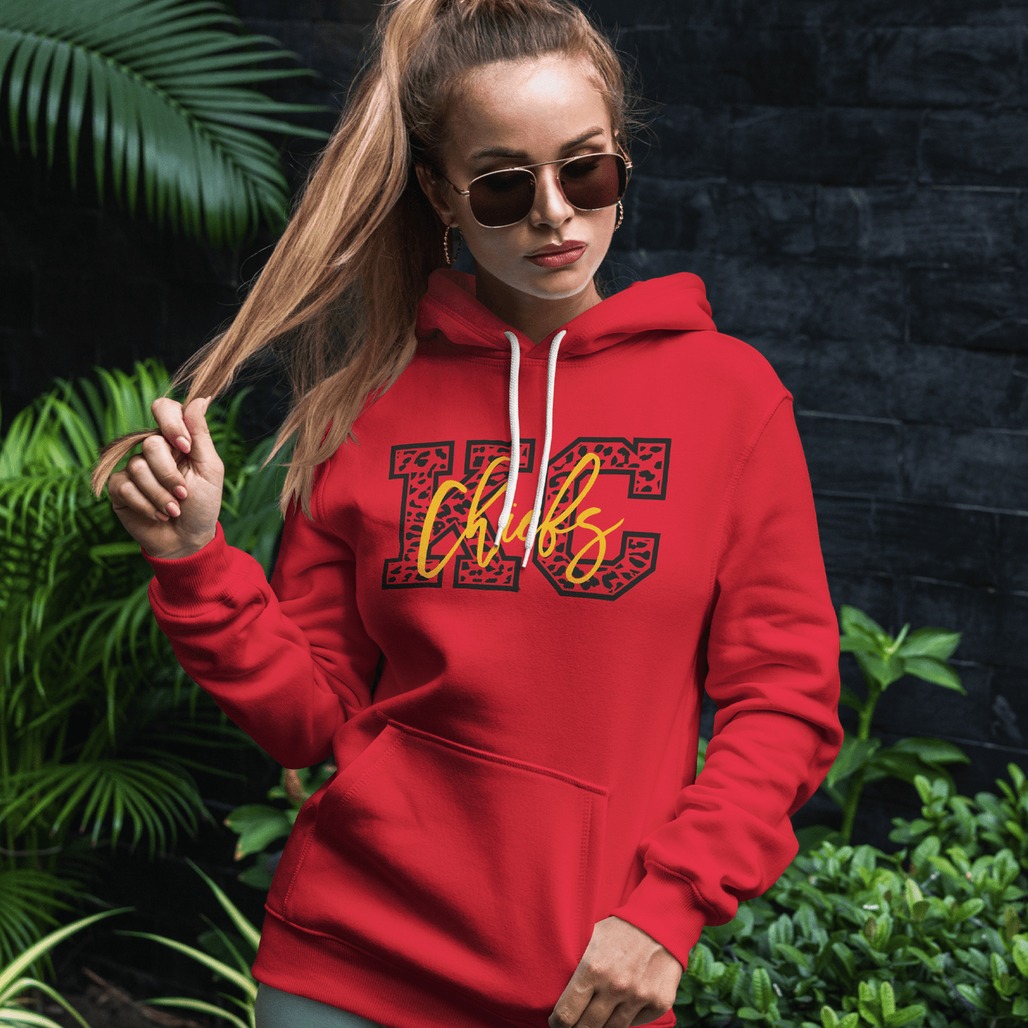 KC Swag Kansas City Chiefs yellow CHIEFS on black cheetah pattern KC on red fleece pullover hoodie worn by female model in front of dark grey wall and green plants