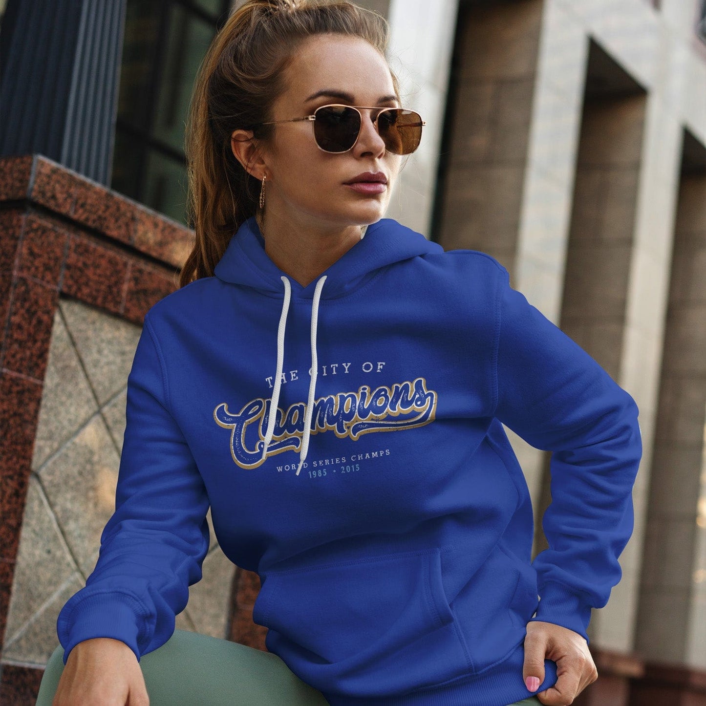 KC Swag Kansas City Royals white/gold CITY OF CHAMPIONS on royal blue pull-over hoodie worn by female model sitting on steps in front of downtown building