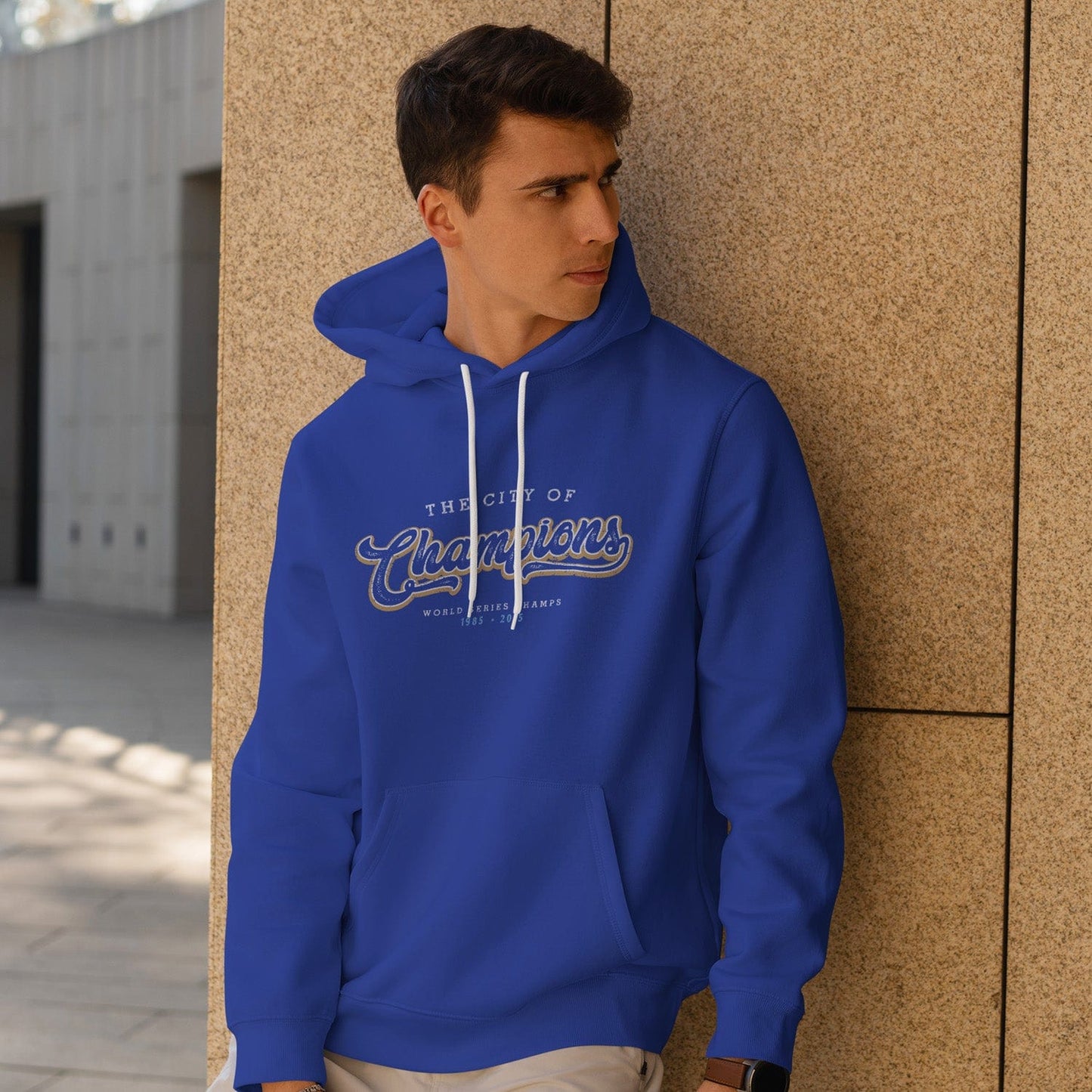 KC Swag Kansas City Royals white/gold CITY OF CHAMPIONS on royal blue pull-over hoodie worn by male model leaning against stone wall in outdoor plaza