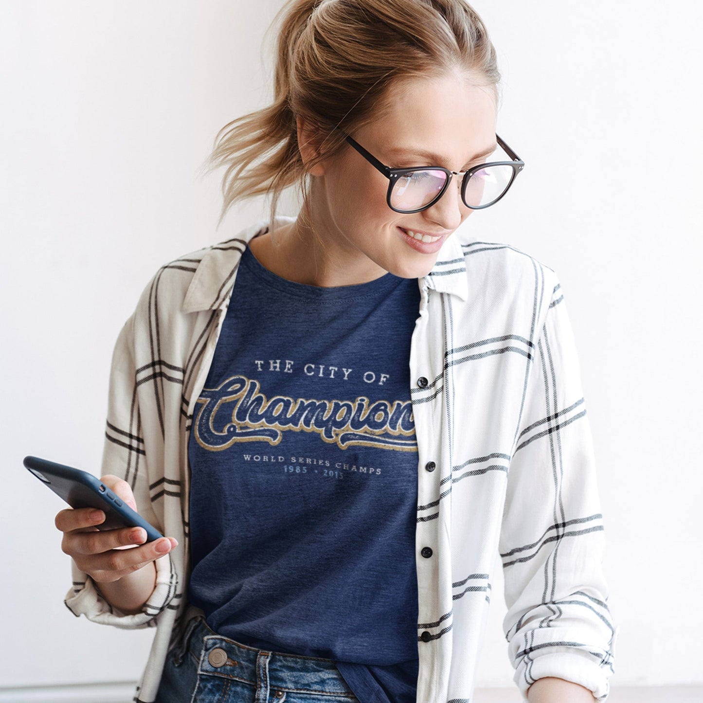 KC Swag Kansas City Royals white/gold CITY OF CHAMPIONS on heather navy t-shirt worn by female model under an open white plaid shirt holding phone
