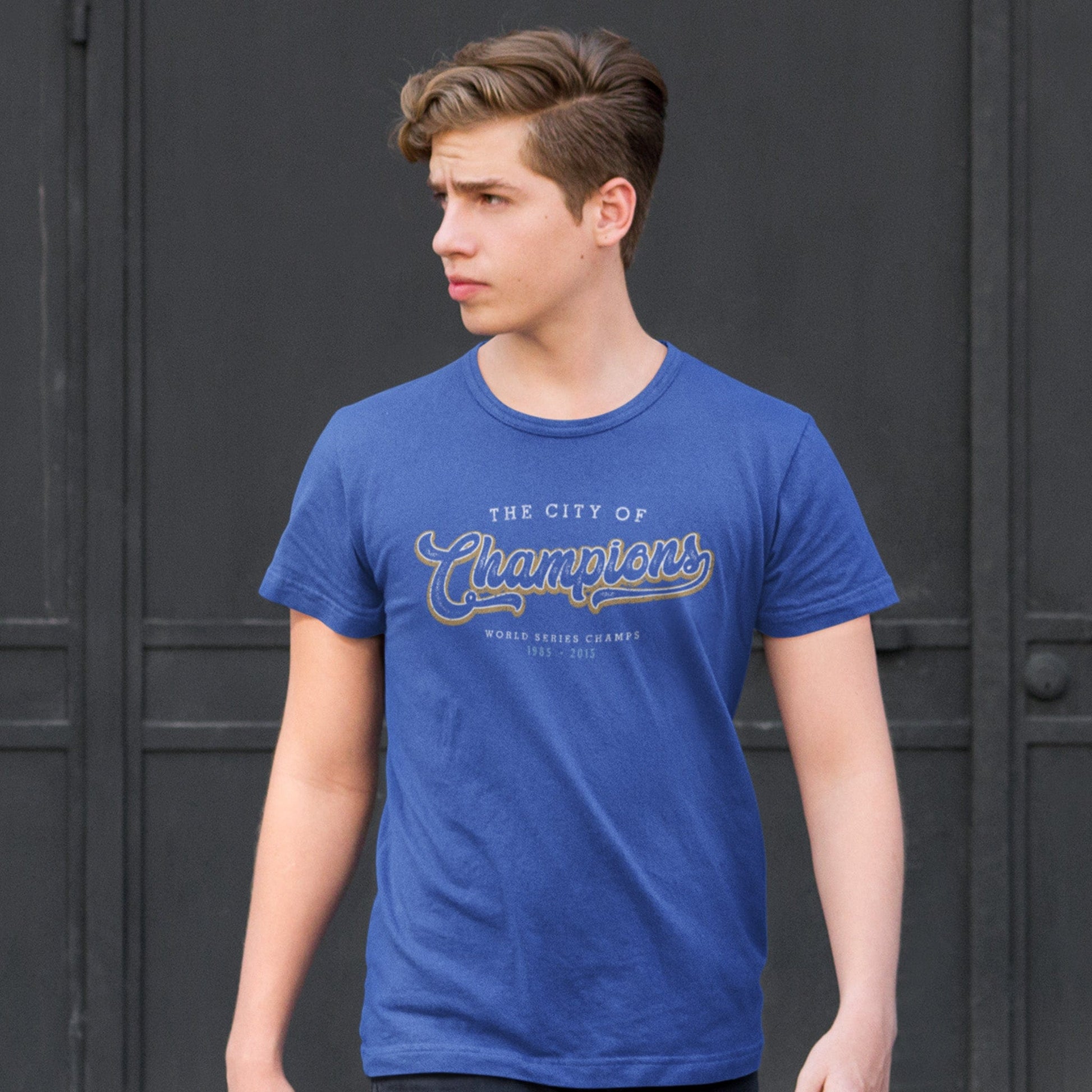 KC Swag Kansas City Royals white/gold CITY OF CHAMPIONS on heather blue t-shirt worn by male model standing in front of large black doors